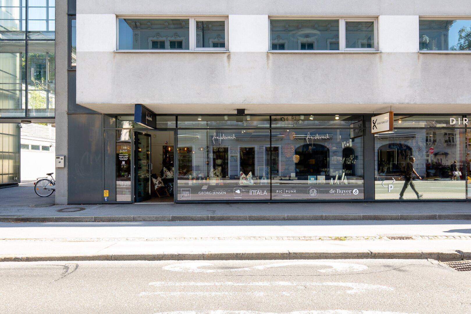Nahaufnahme der Geschäftsfronten mit großen Schaufenstern und Eingangsbereichen an einer belebten Straße.