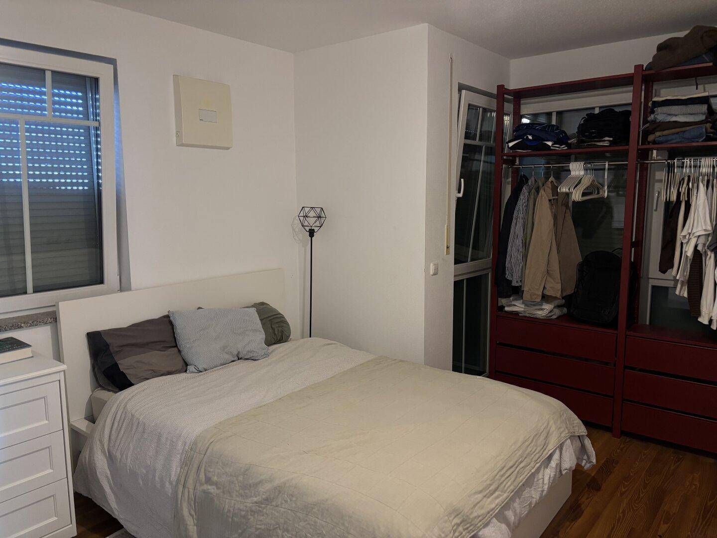 Schlafzimmer mit Doppelbett, Holzboden und offenem Kleiderschrank neben dem Fenster.