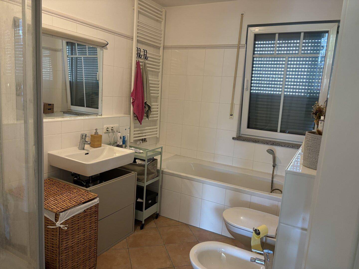Helles Badezimmer mit Badewanne, Dusche, Bidet und Fenster mit weißem Fliesenboden.