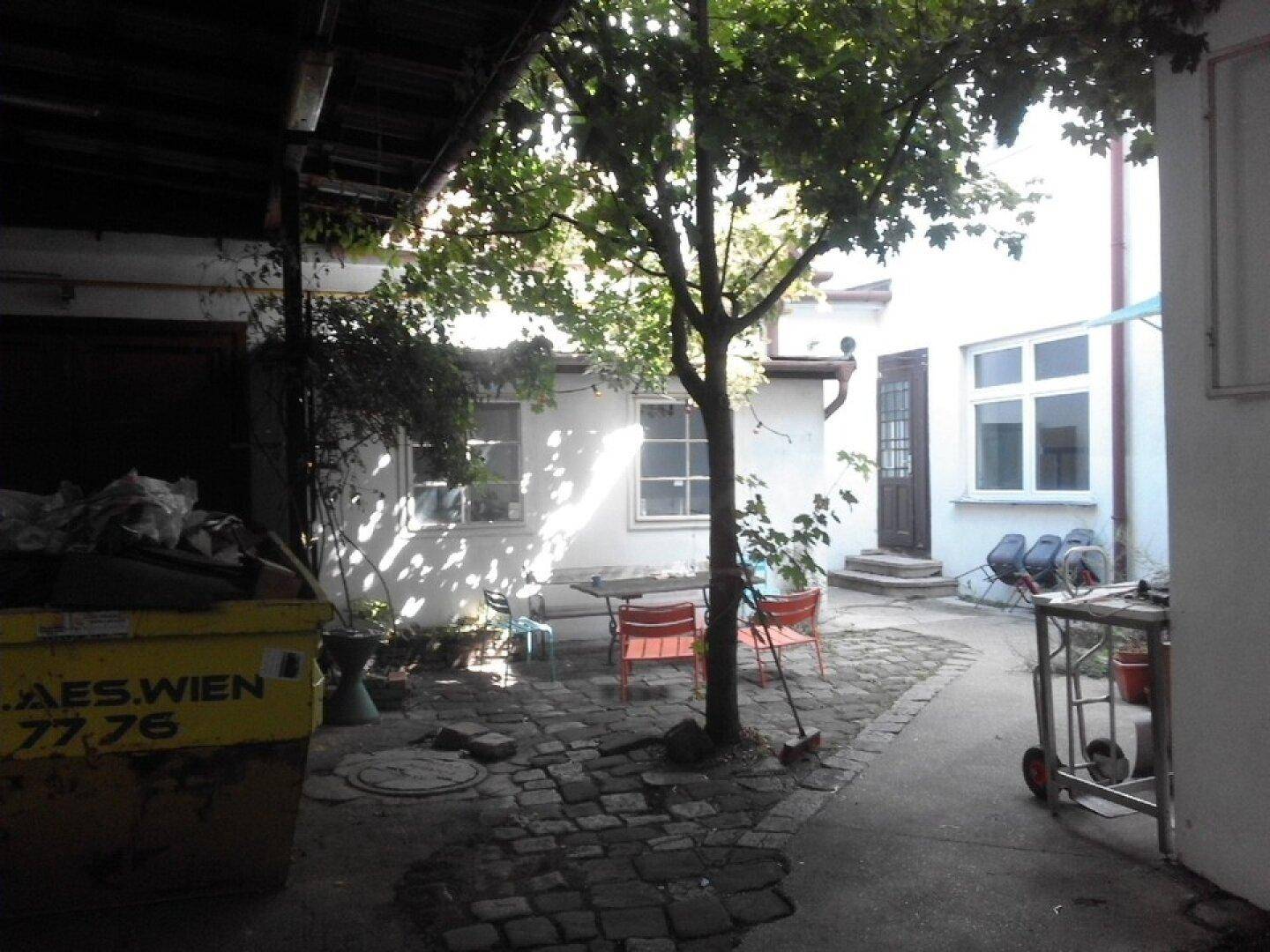 Blick auf einen gepflasterten Innenhof mit einem Baum, Sitzgelegenheiten und einem Müllcontainer.