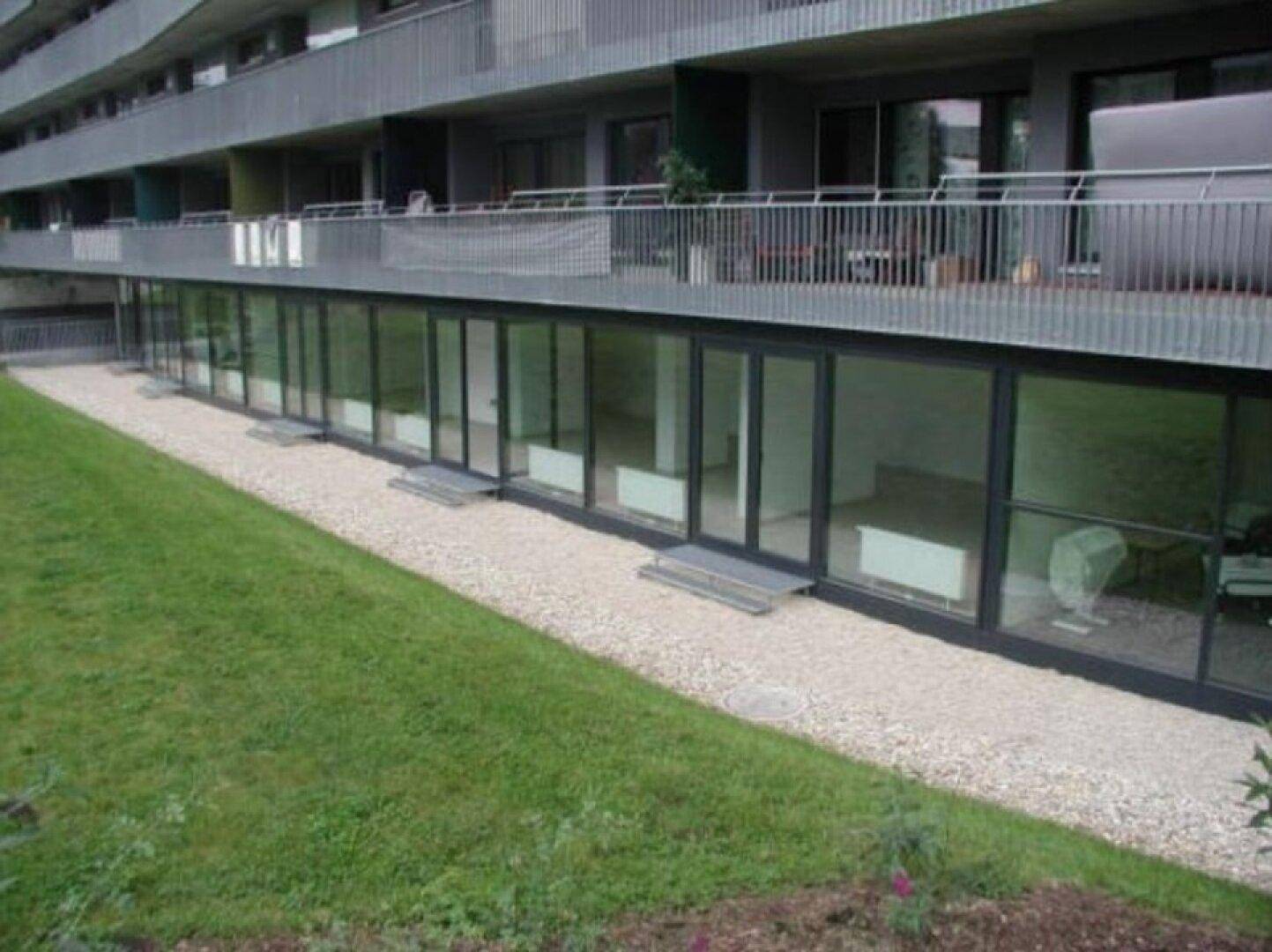 Außenansicht eines modernen Gebäudes mit großen Glasfronten im Erdgeschoss und Balkonen darüber.