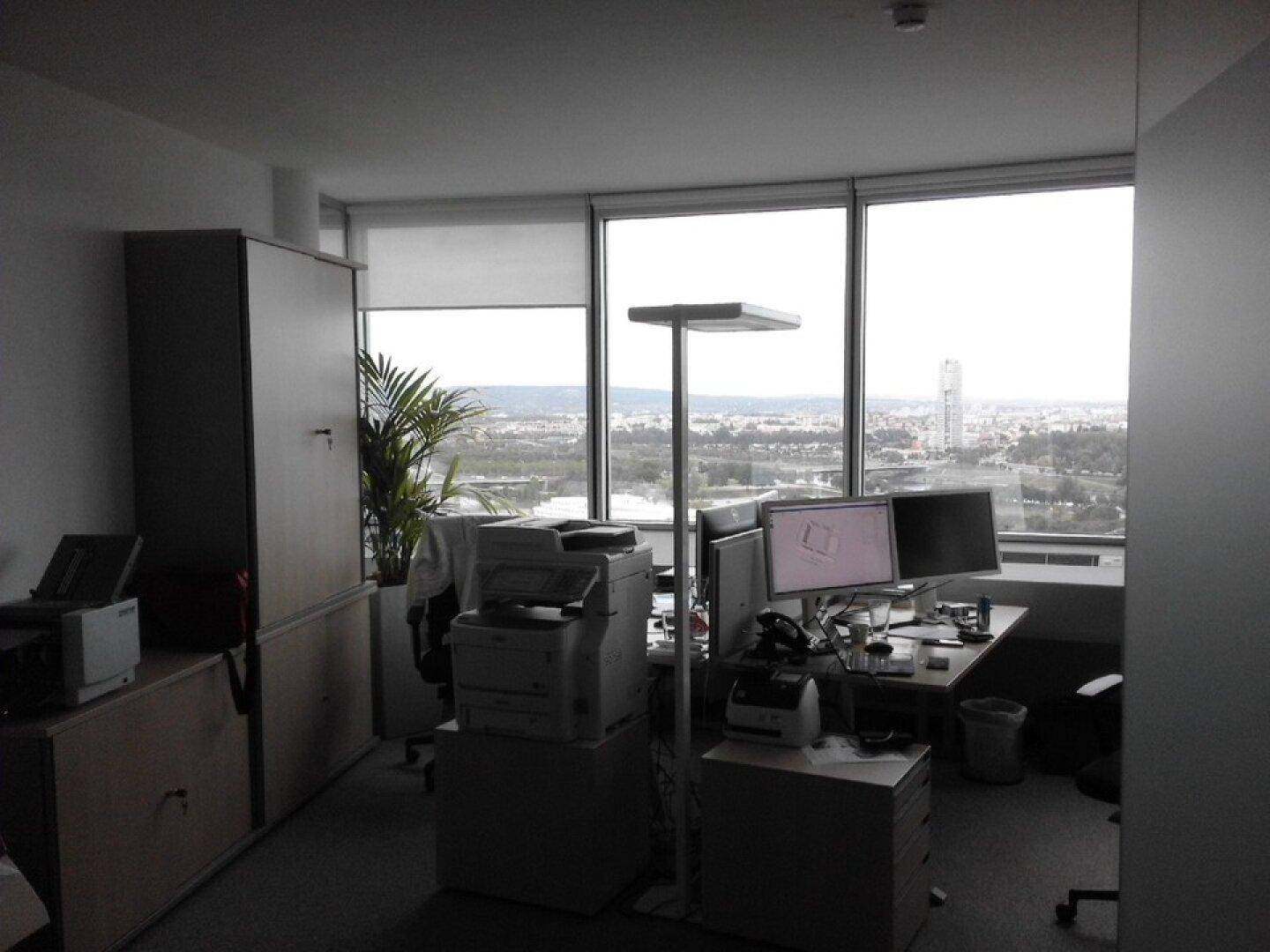 Büroarbeitsplatz mit Schreibtisch, Computern, Drucker und weitem Blick auf die Stadt durch große Fenster.