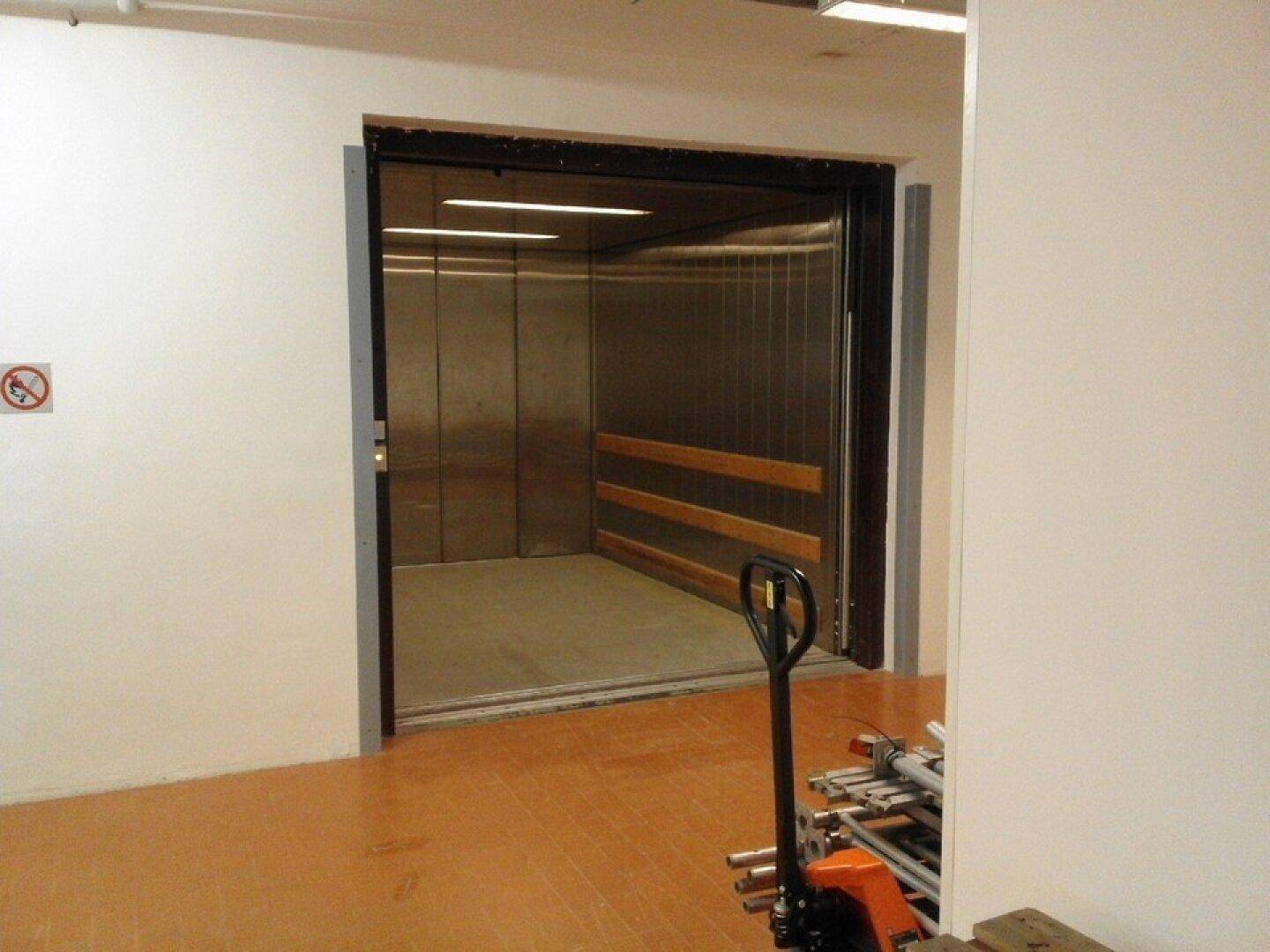 Geöffneter Lastenaufzug mit Metallwänden und Holzschutzleisten, bereit für den Transport von Gütern.