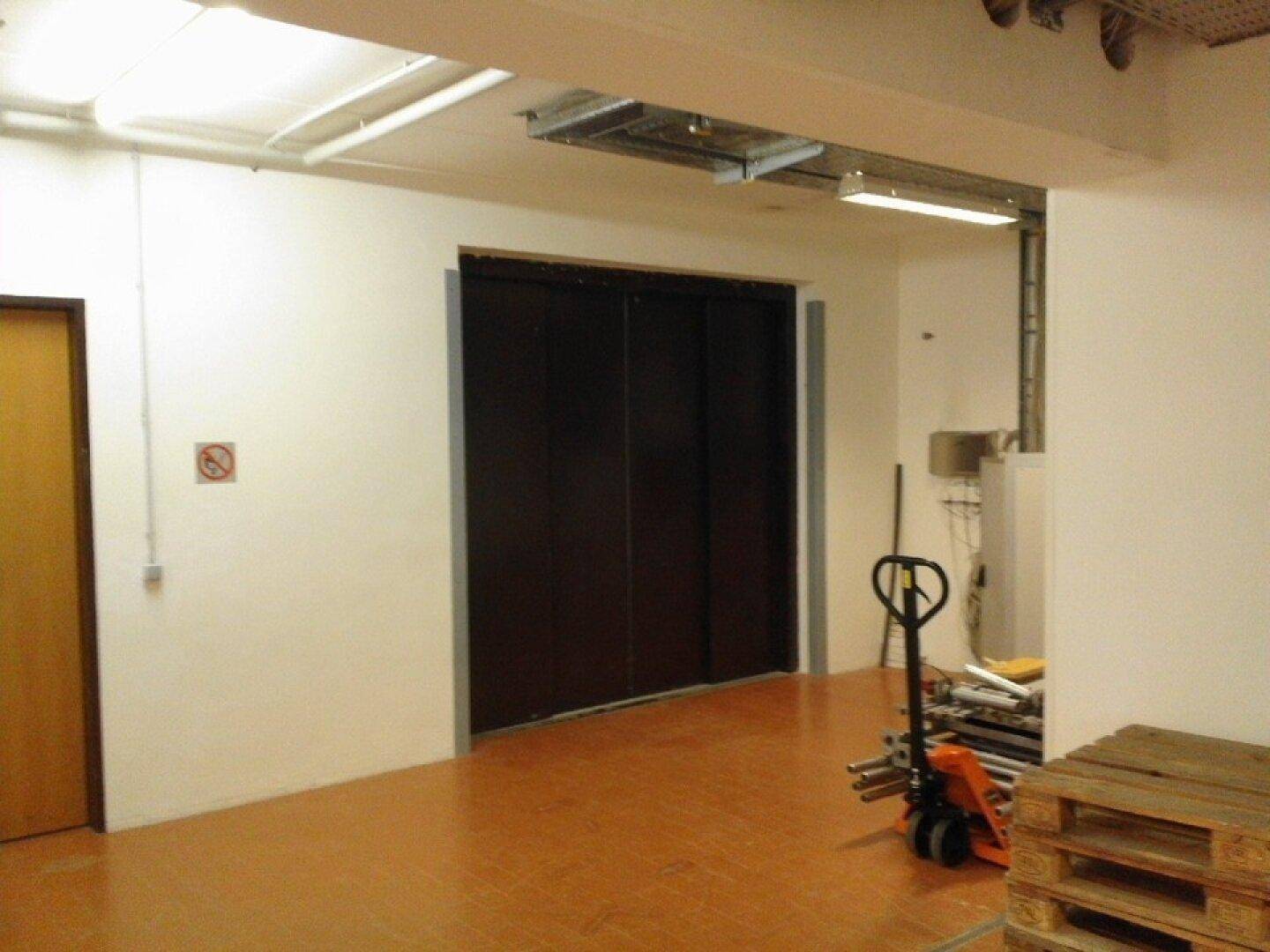 Geschlossene Aufzugstüren in einem Flur, daneben ein Hubwagen und Holzpaletten, was auf Lagerung hindeutet.