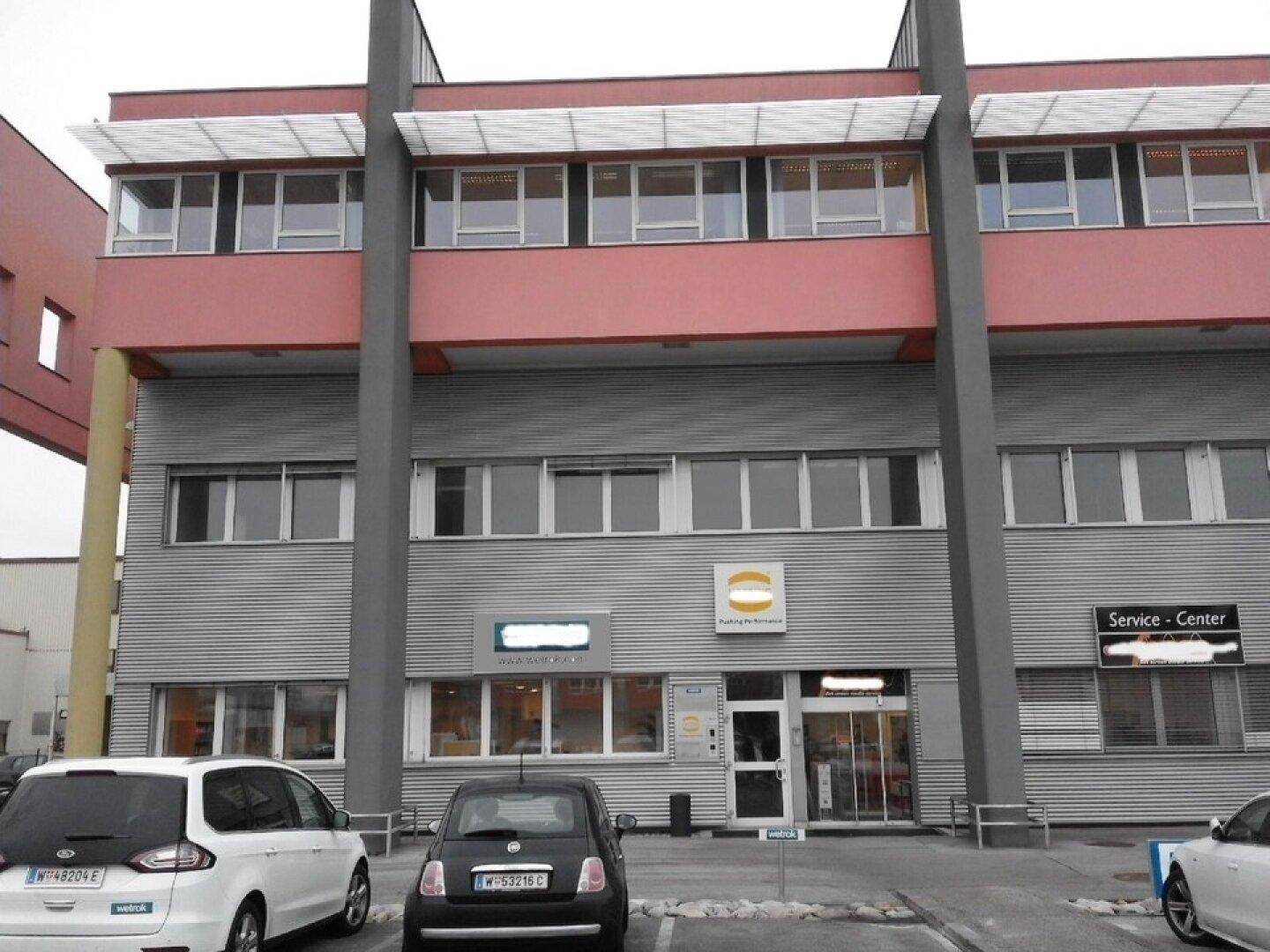 Modernes Geschäftsgebäude mit roter und grauer Fassade sowie Parkplätzen davor.
