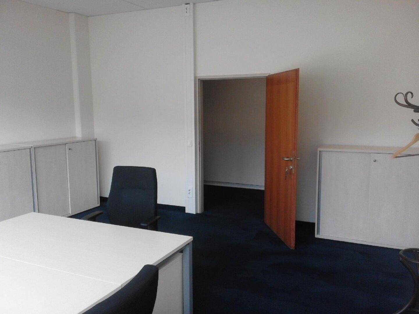 Büro mit weißem Mobiliar, Teppichboden und offener Tür zu einem weiteren Raum, bietet flexible Nutzungsmöglichkeiten.