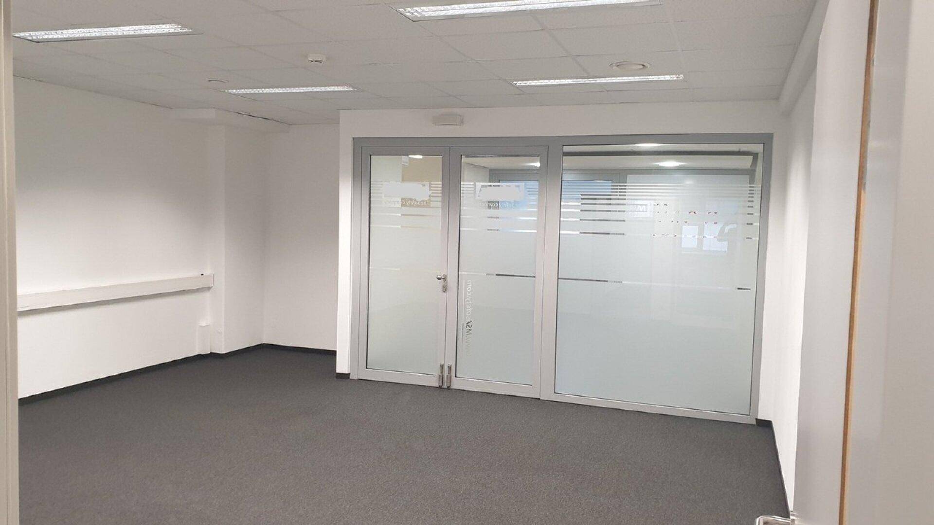 Leerer Büroraum mit Teppichboden und einer modernen Glaswand, die einen weiteren Bereich abtrennt.