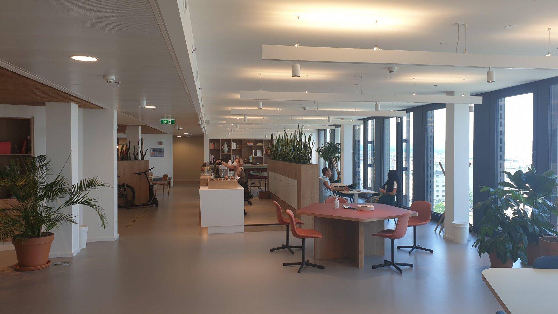 Geräumiger offener Bürobereich mit verschiedenen Sitzgelegenheiten, Pflanzen und großen Fenstern.