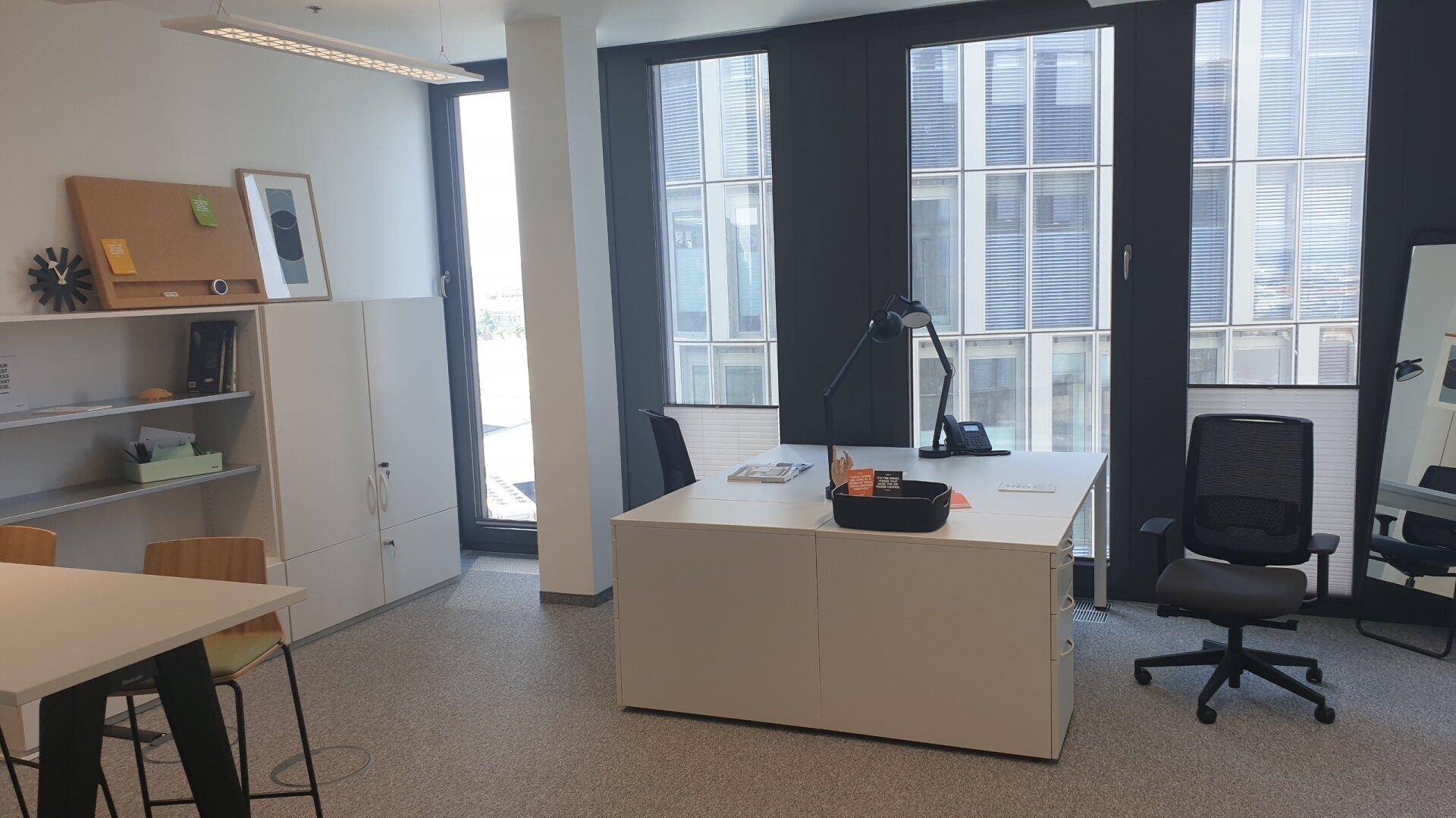 Geräumiges Büro mit einem Schreibtisch, Bürostuhl und hohen Fenstern, die viel Tageslicht hereinlassen.
