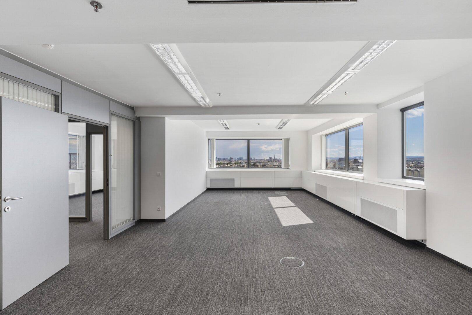 Geräumiges, leeres Büro mit Teppichboden, großen Fenstern und weitem Blick auf die Stadt.