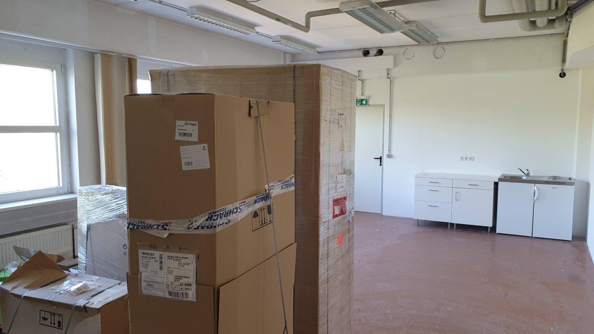 Raum mit einer kleinen Küchenzeile, verpackten Kartons auf Paletten und Fenstern, die Tageslicht hereinlassen.