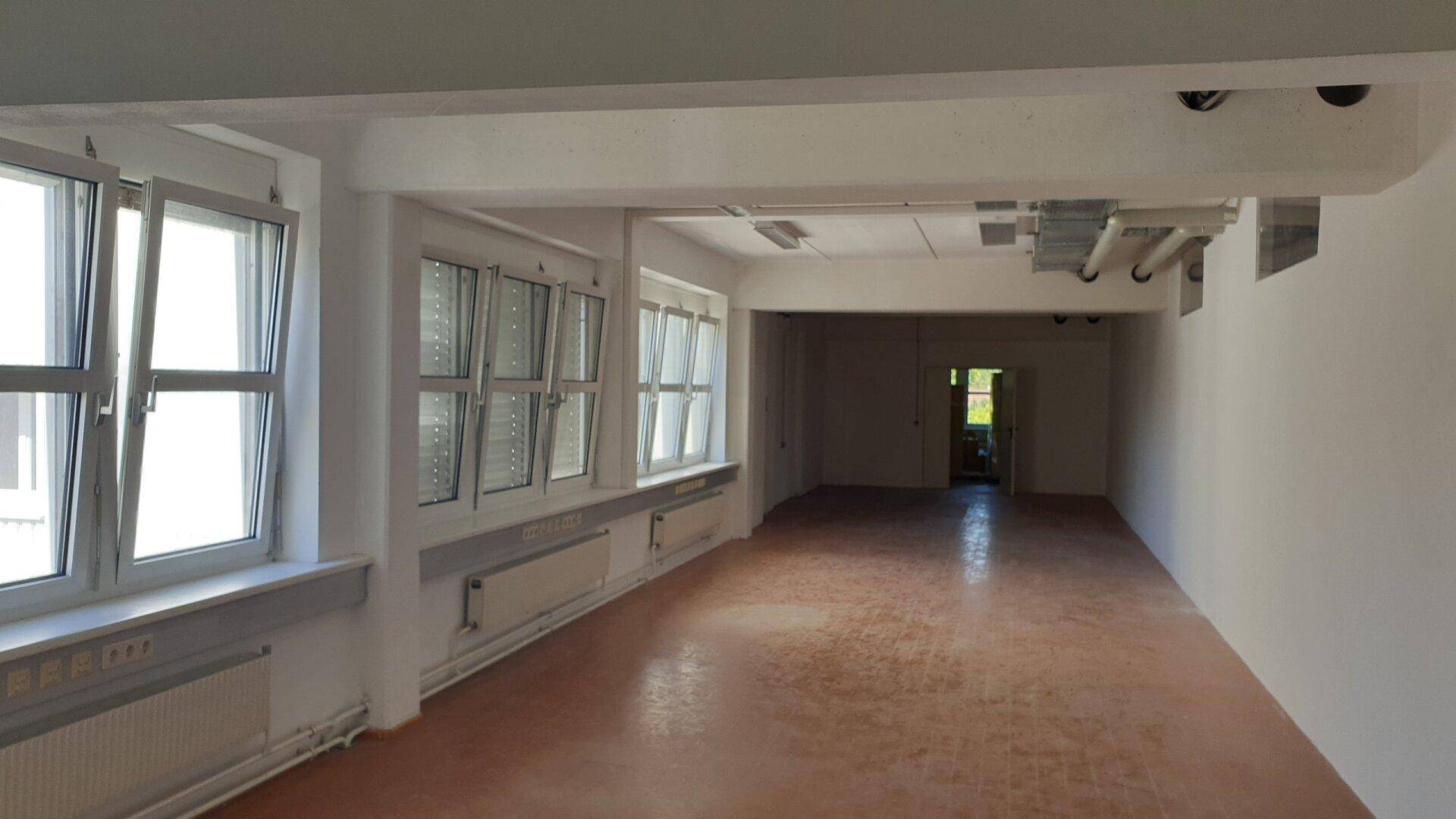 Geräumiger, leerer Raum mit einer Reihe von Fenstern, sichtbaren Deckeninstallationen und einem funktionalen Boden.