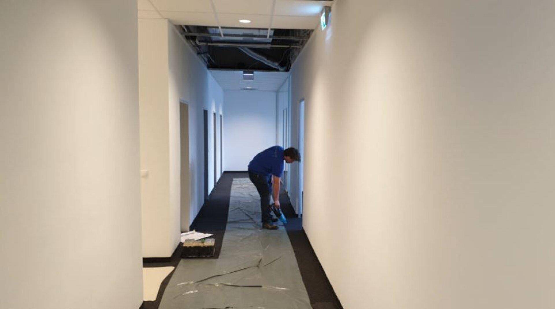 Langer Flur mit Teppichboden und teilweise offener Decke, wo ein Arbeiter Reinigungsarbeiten durchführt.