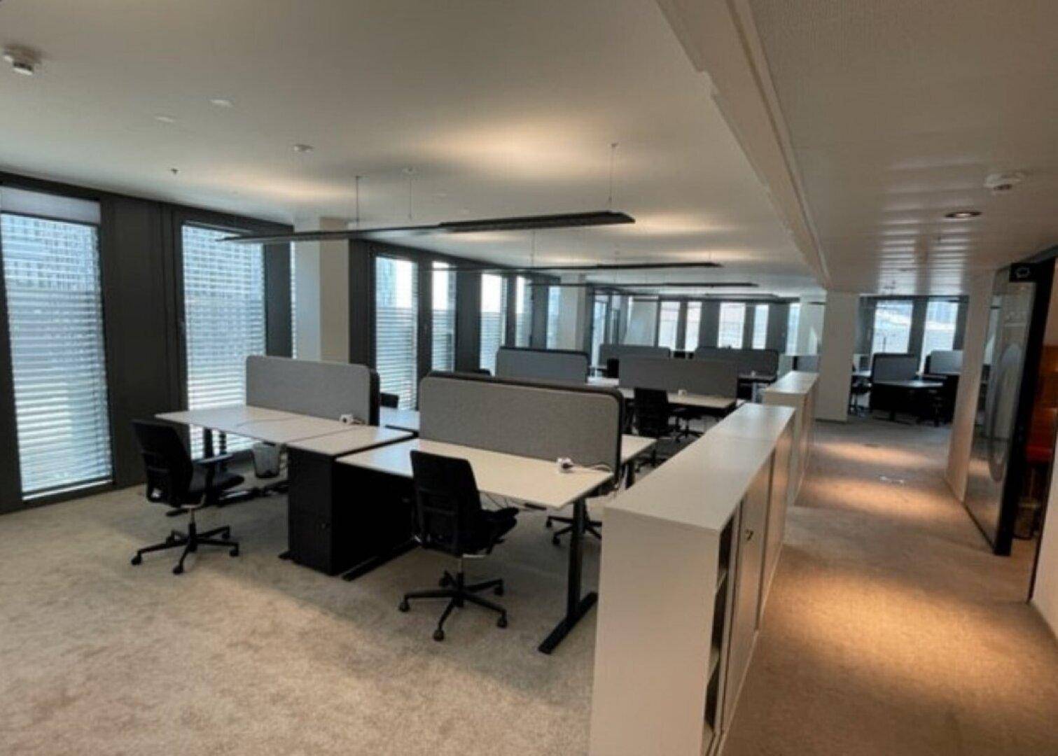 Geräumiges Großraumbüro mit modernen Arbeitsplätzen, Teppichboden und großen Fenstern mit Jalousien.