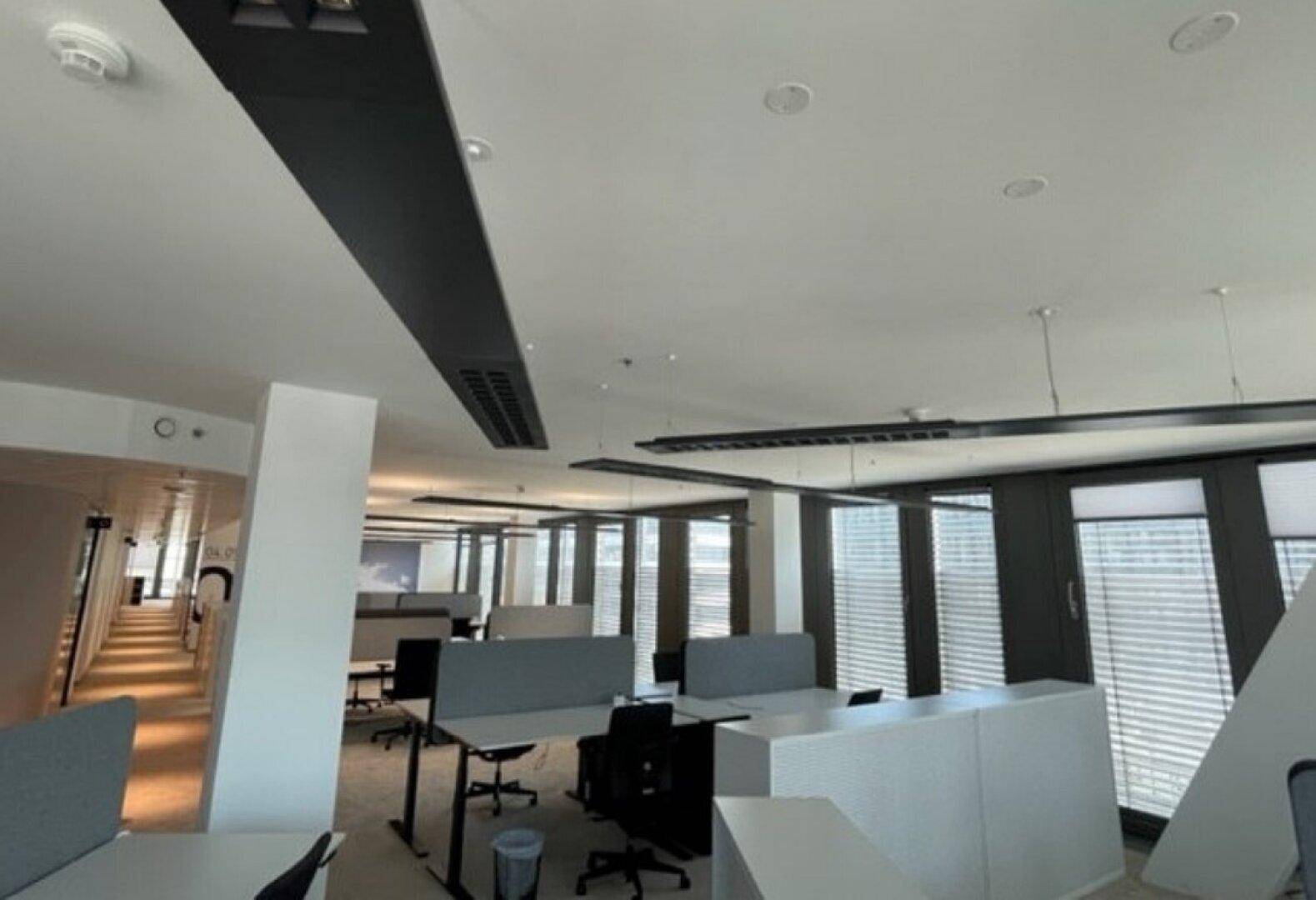 Großraumbüro mit großen Fenstern, Jalousien und moderner Deckenbeleuchtung, die viel Tageslicht hereinlässt.