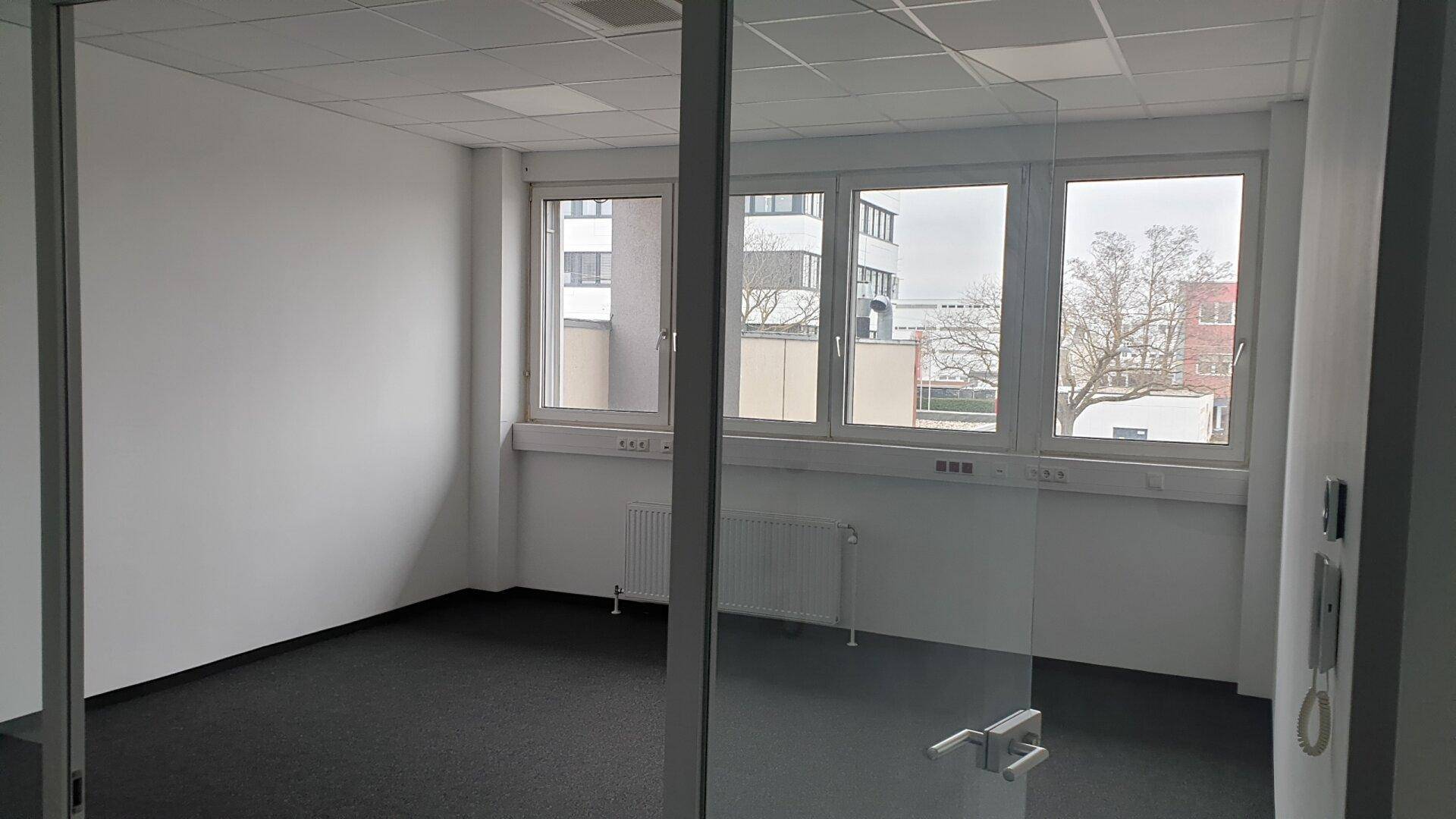 Geräumiges Büro mit Teppichboden, großen Fenstern und Blick auf umliegende Gebäude.