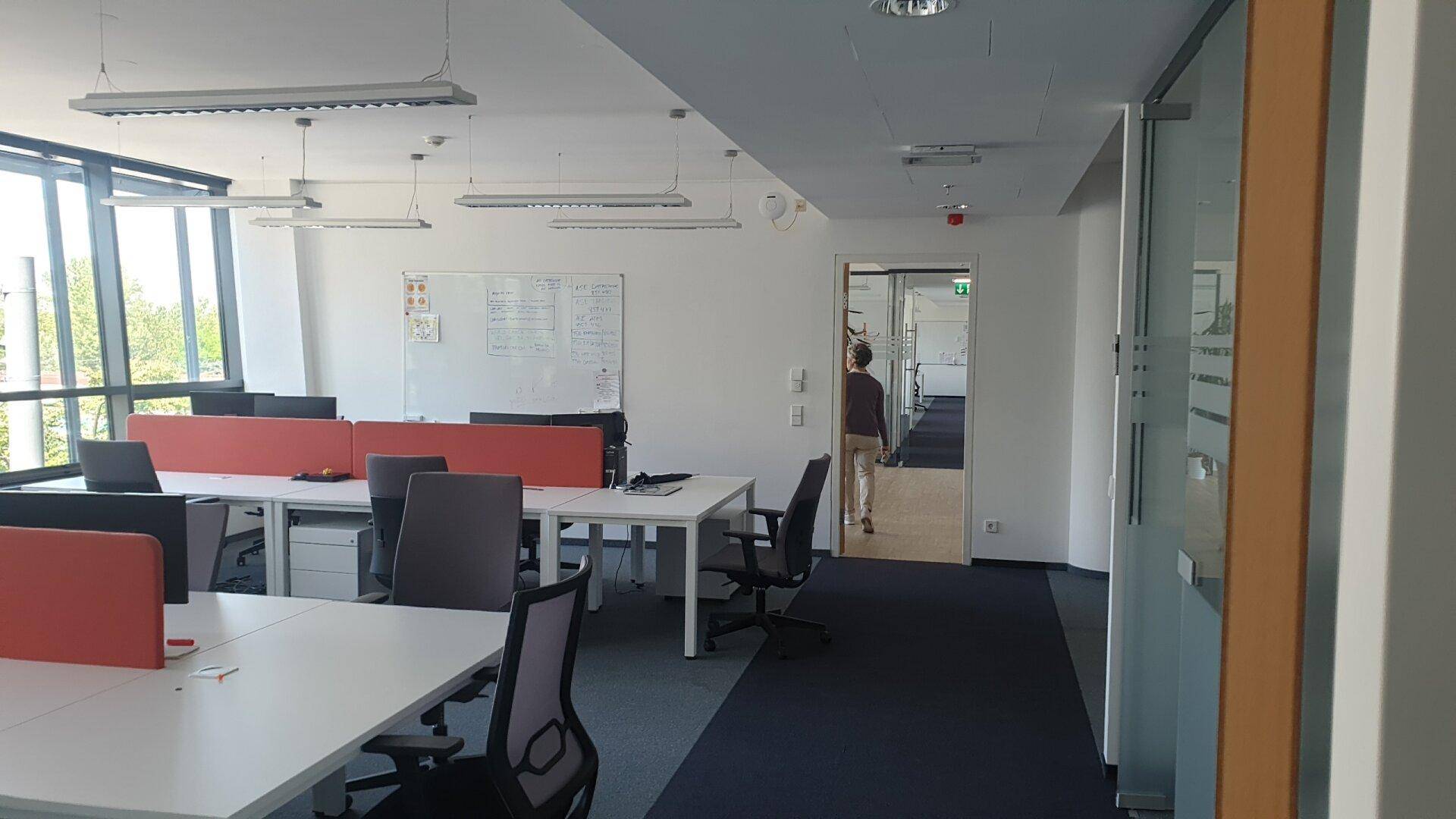 Geräumiges Büro mit vielen Fenstern, Whiteboard und Zugang zu einem Flur.