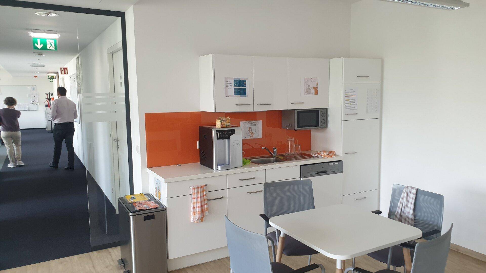 Kleine Teeküche mit weißen Schränken, orangefarbener Rückwand und Essbereich für Mitarbeiter.