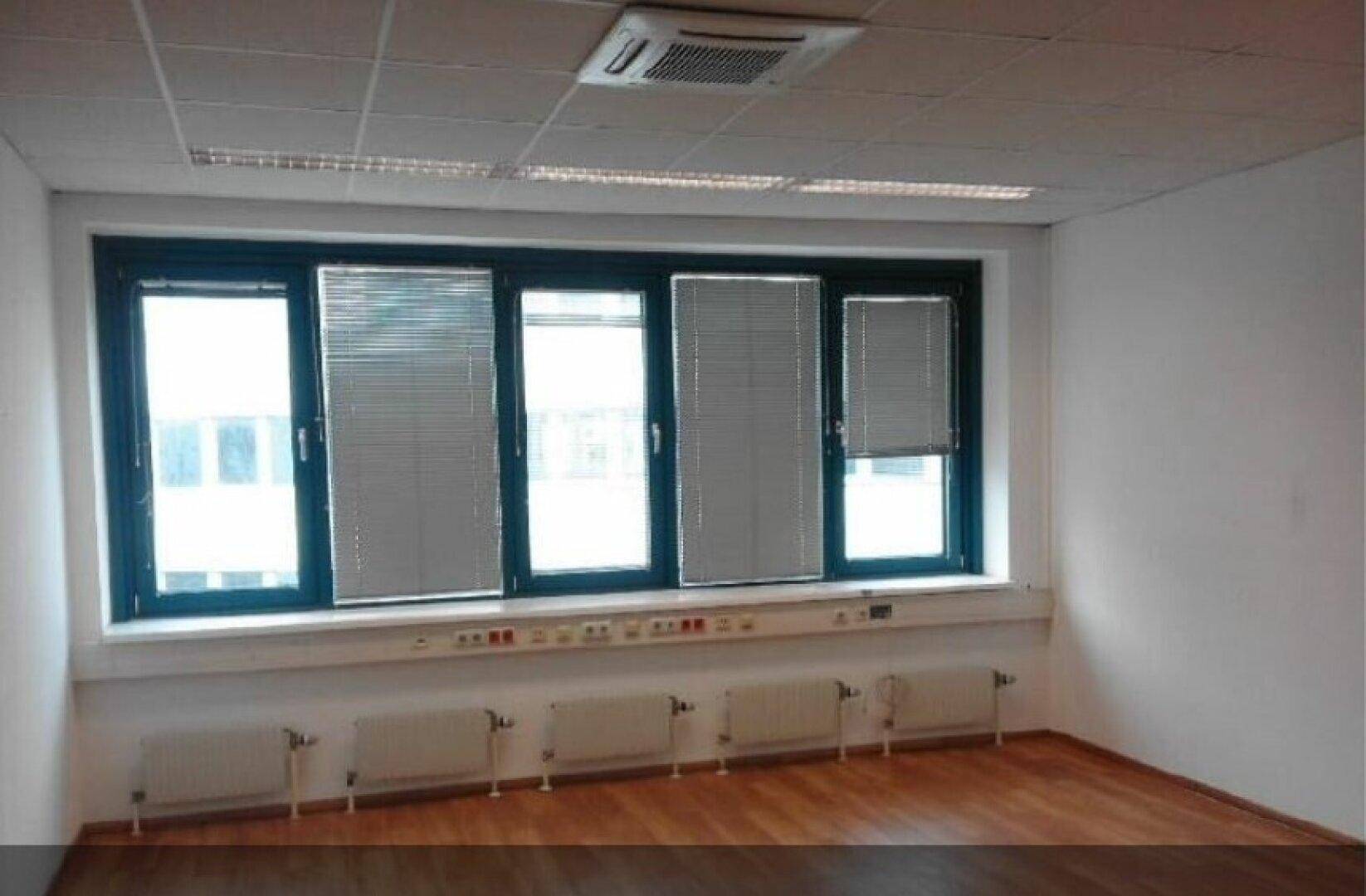 Leeres Büro mit Holzfußboden, großen Fenstern mit Jalousien und Deckenbeleuchtung.