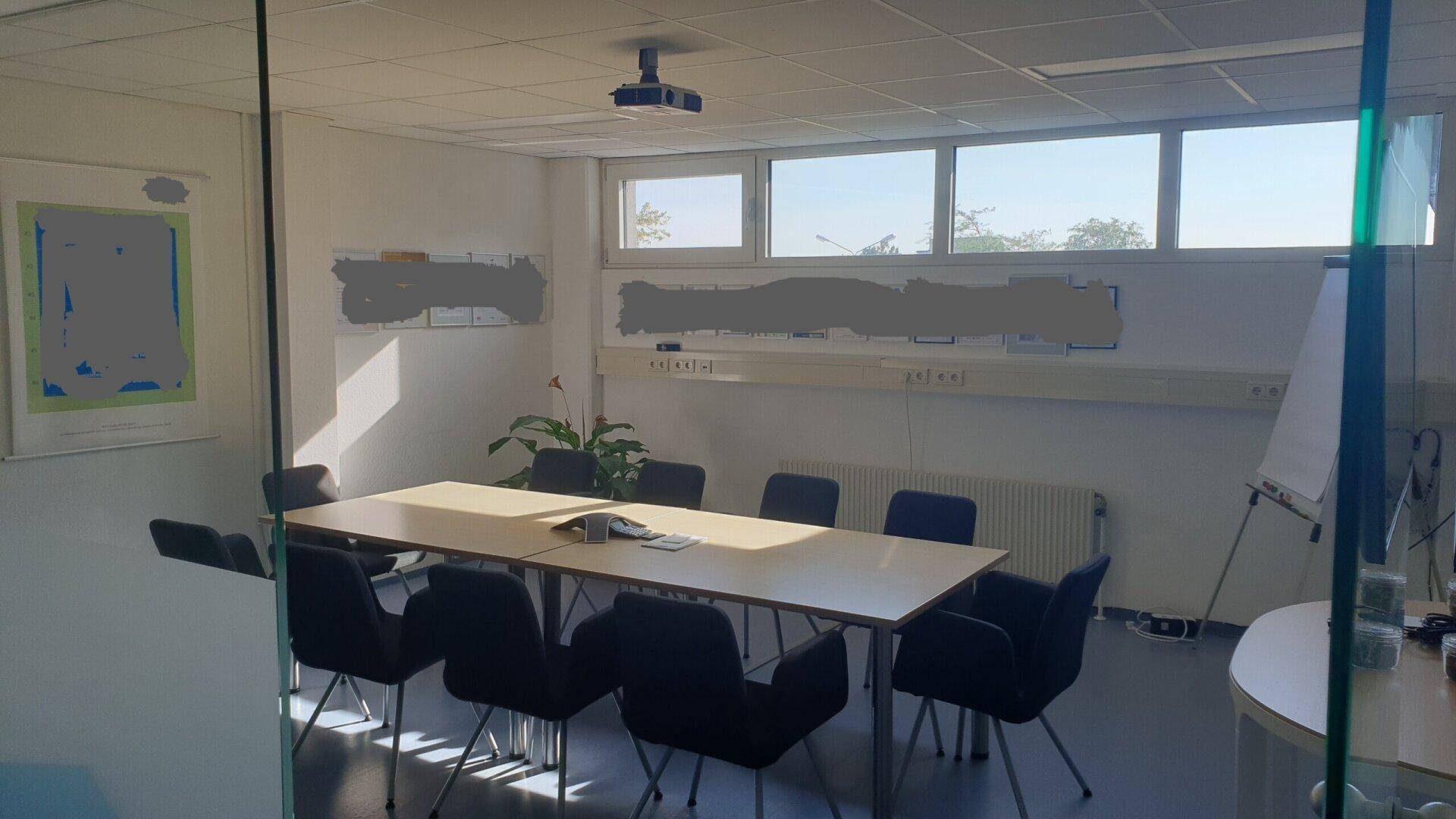 Konferenzraum mit großem Tisch, Stühlen, Beamer und Fenstern für Tageslicht.