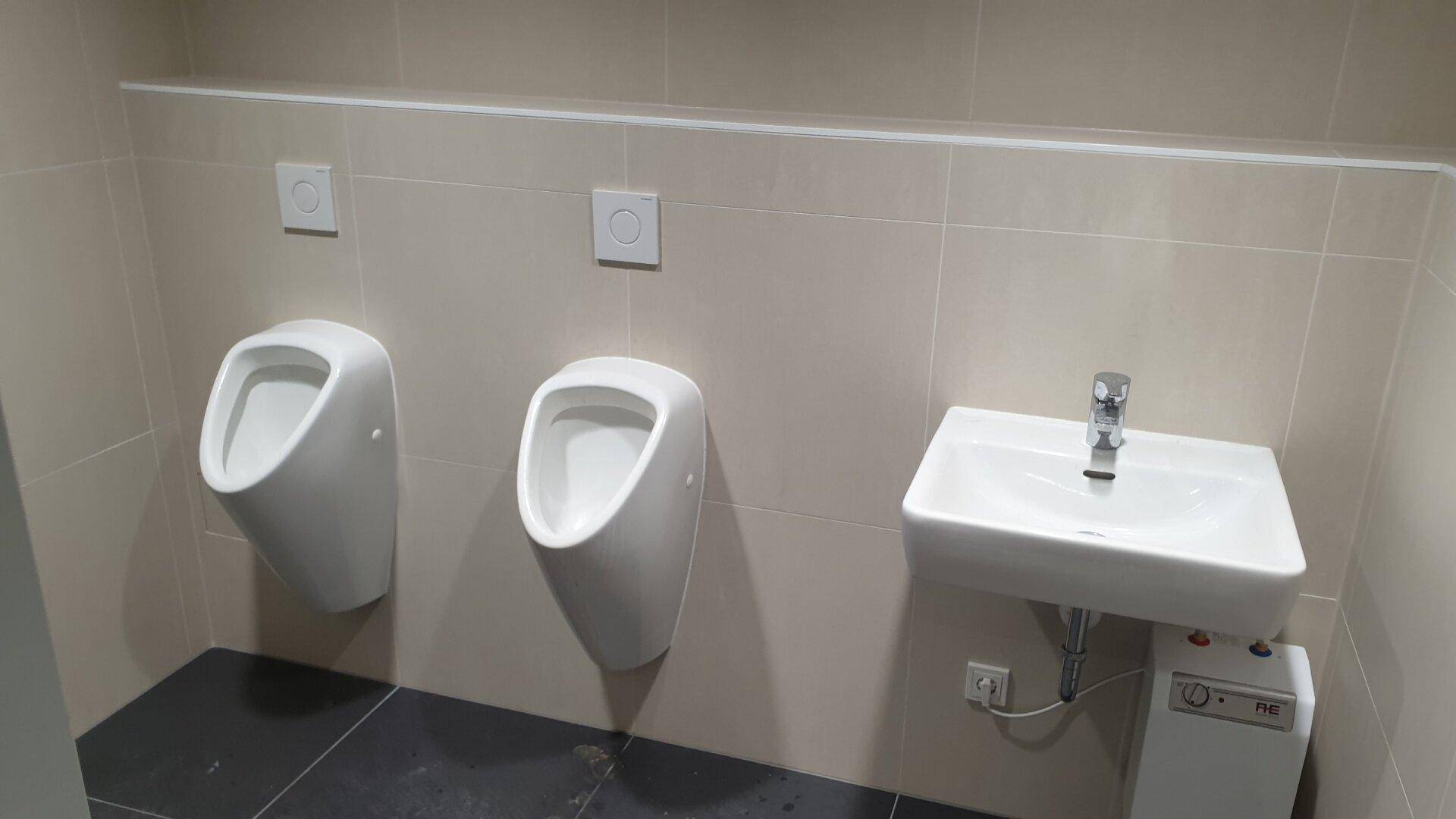 Modern geflieste Herrentoilette mit zwei Urinalen und einem Waschbecken.