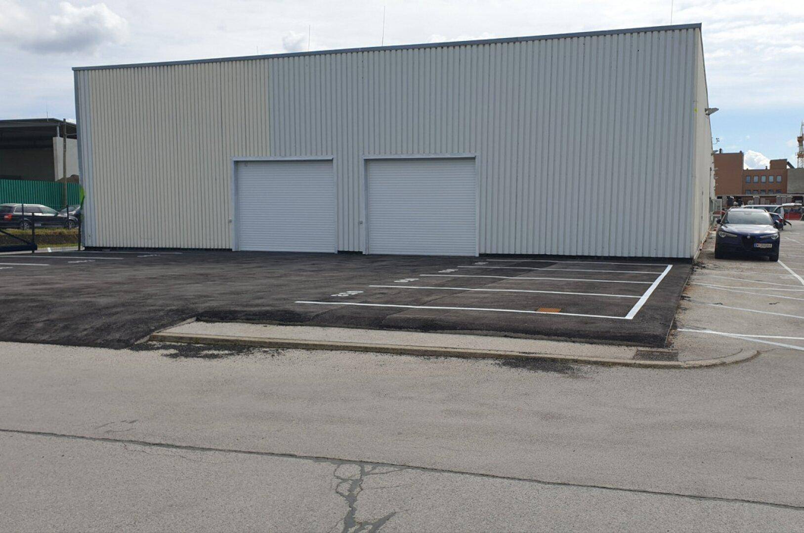 Modernes Industriegebäude mit zwei Rolltoren und asphaltiertem Parkplatz, umgeben von weiteren Gebäuden.
