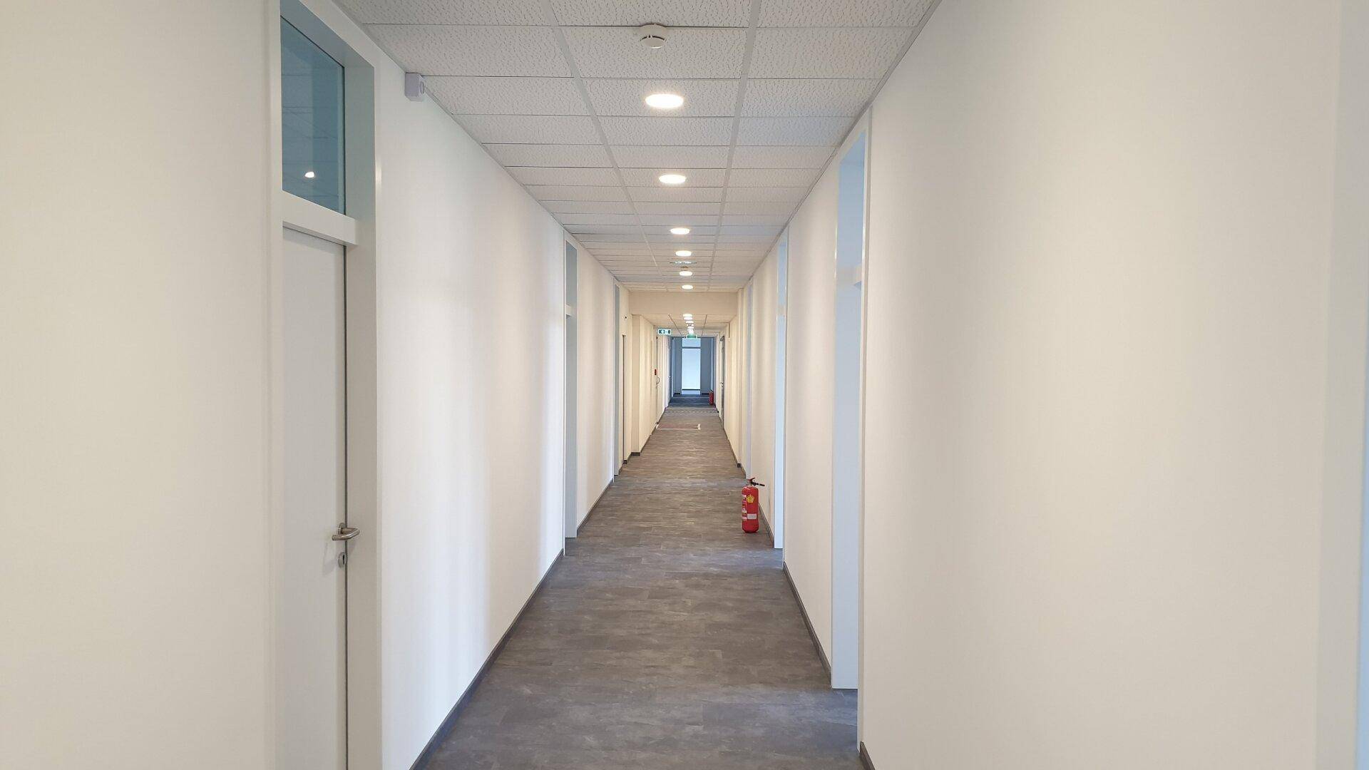 Langer, heller Flur mit weißen Wänden, abgehängter Decke und grauen Bodenfliesen, führt zu weiteren Räumen.