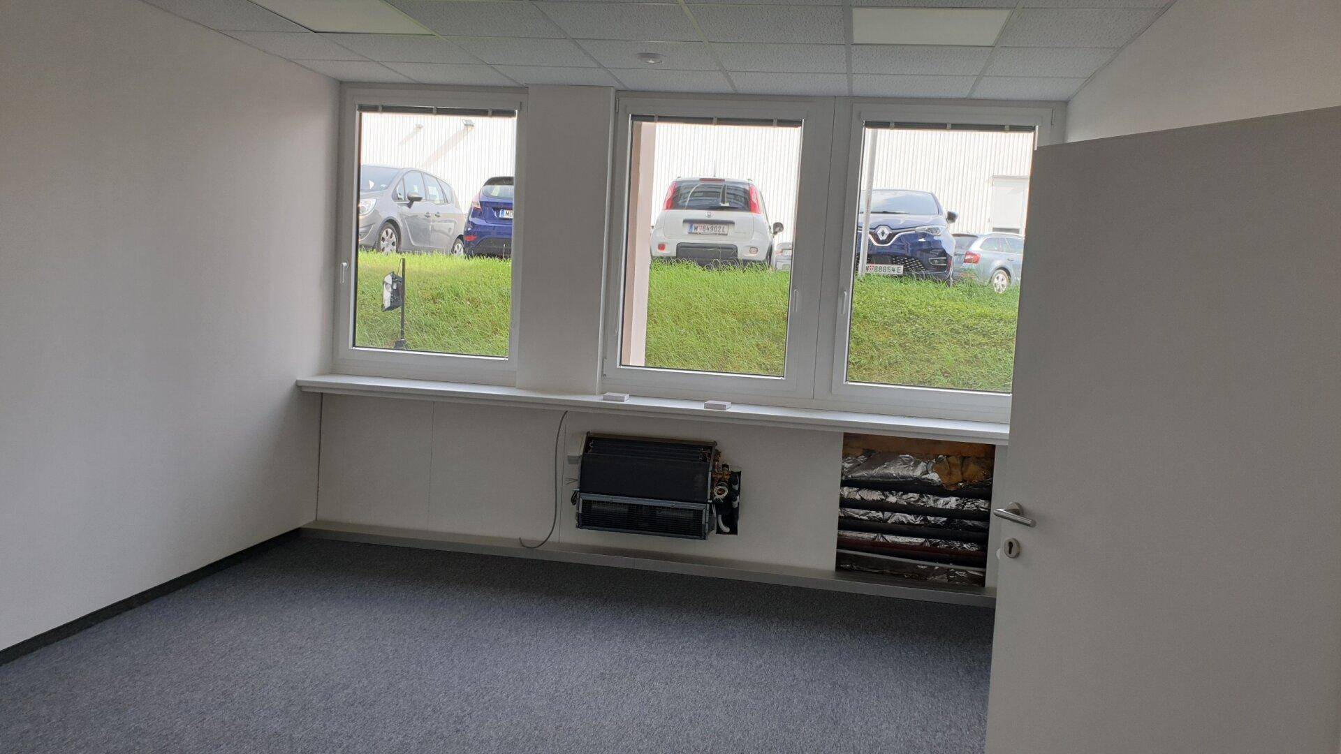 Leeres Büro mit Teppichboden, großen Fenstern und Blick auf den Parkplatz, bereit zur individuellen Gestaltung.