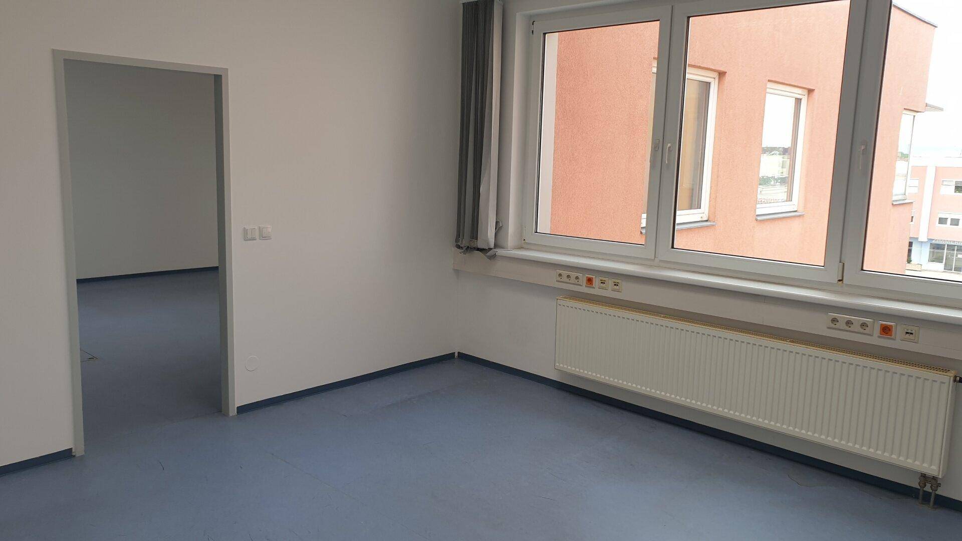 Heller Raum mit großem Fenster, blauem Boden und Blick auf ein gegenüberliegendes Gebäude.