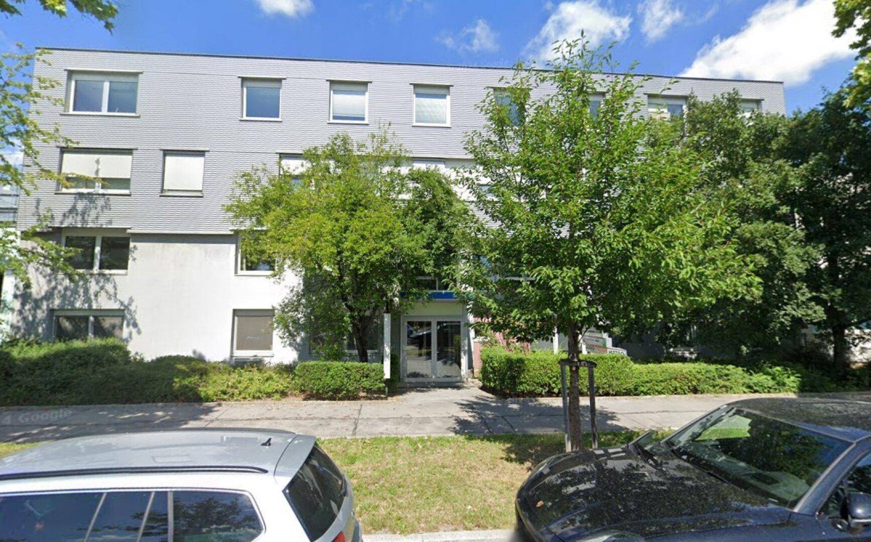 Mehrstöckiges Gebäude mit grauer Fassade, umgeben von Bäumen und Büschen, mit Parkplätzen davor.