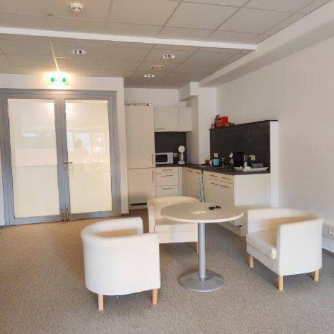 Moderne Teeküche in einem Büro mit hellen Schränken, Sitzgelegenheiten und Teppichboden.