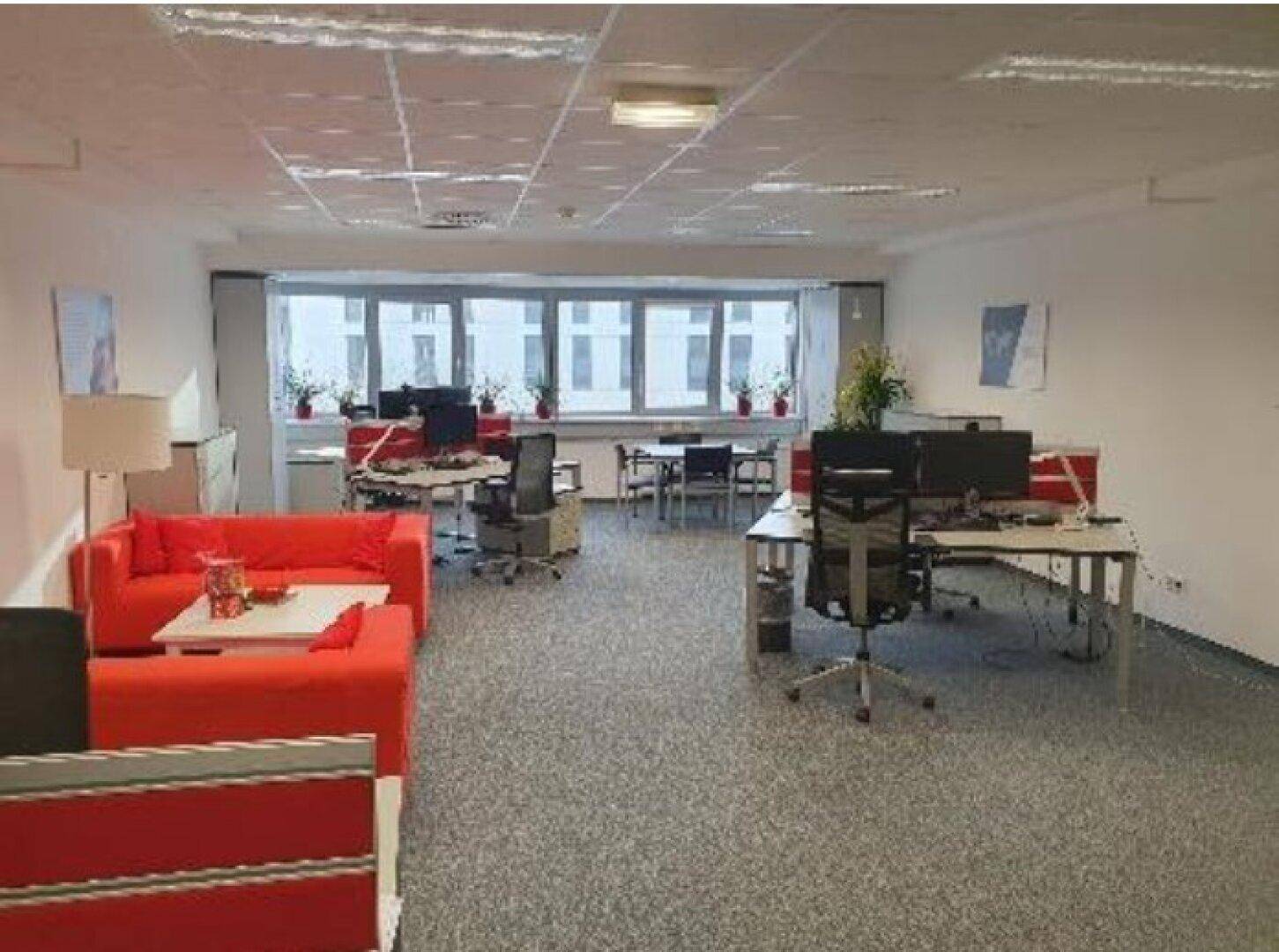 Großzügiges Großraumbüro mit roten Sofas, mehreren Arbeitsplätzen und Blick nach draußen.