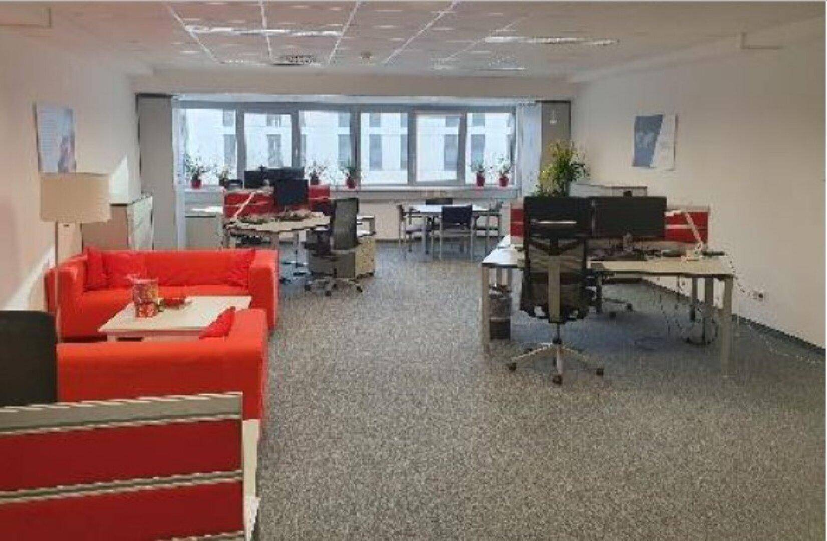 Großzügiges Großraumbüro mit roten Sofas, mehreren Arbeitsplätzen und Blick nach draußen.