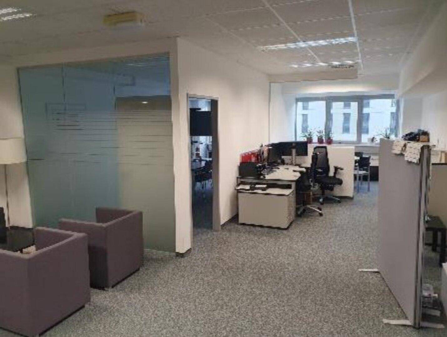 Offener Bürobereich mit Teppichboden, Arbeitsplätzen und einer modernen Glaswand als Raumteiler.