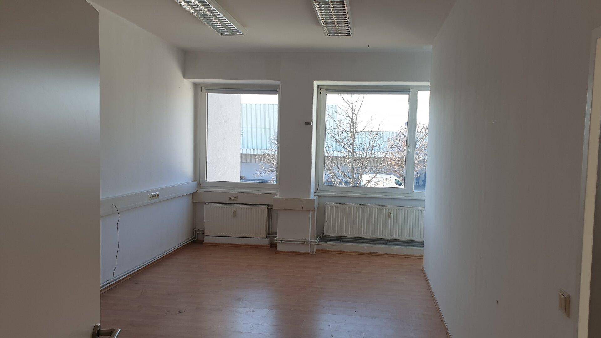 Heller Büroraum mit zwei großen Fenstern, Laminatboden und Heizkörpern, bietet viel Tageslicht.