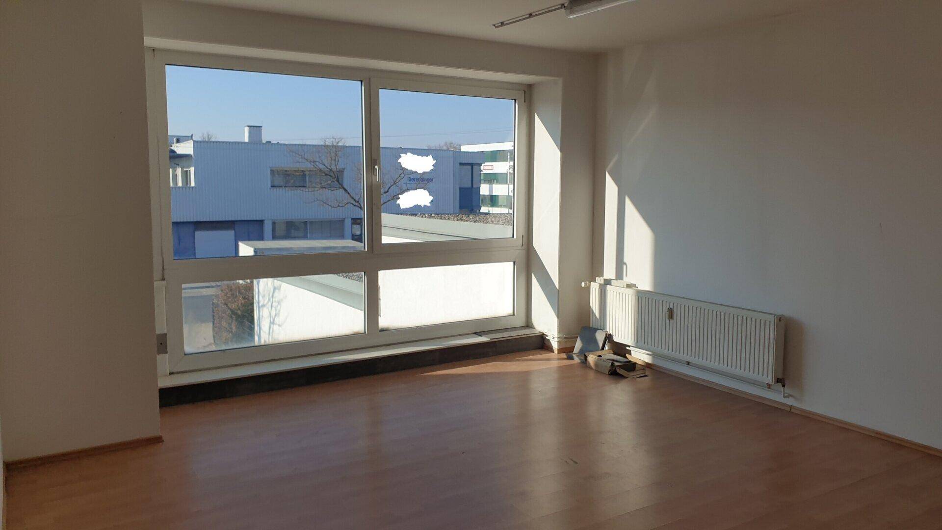 Sonniger Büroraum mit großem Fenster, Laminatboden und Heizkörper, bietet weite Aussicht und viel Helligkeit.