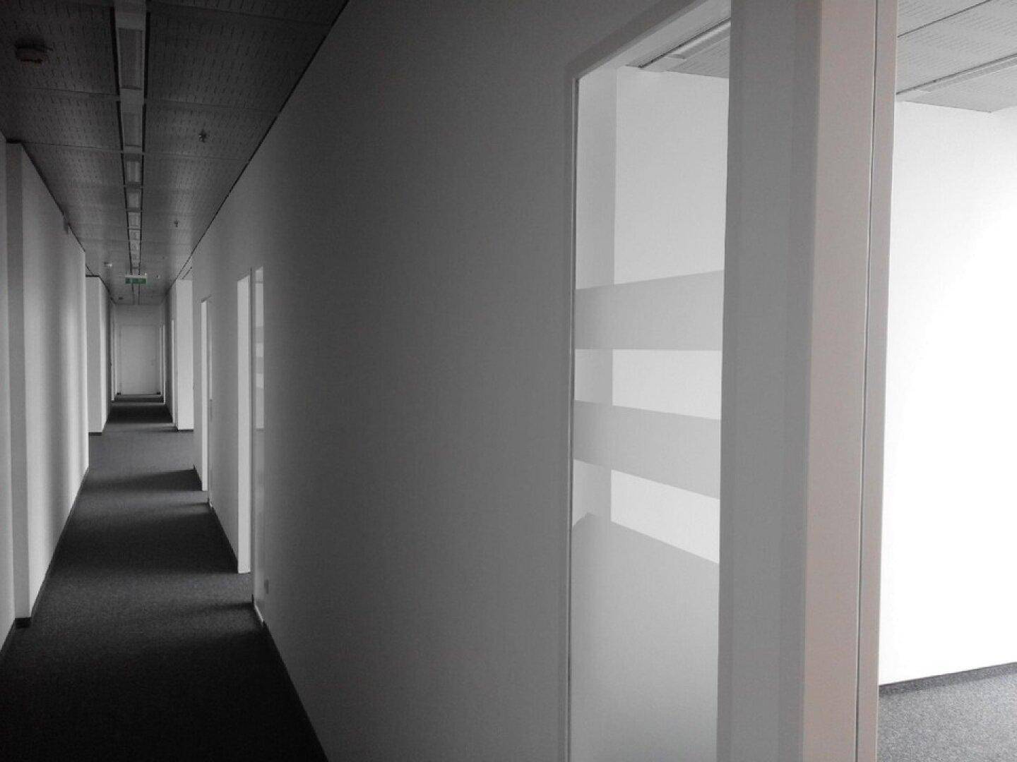 Langer, heller Büroflur mit weißen Wänden und dunklem Teppichboden, der zu weiteren Räumen führt.