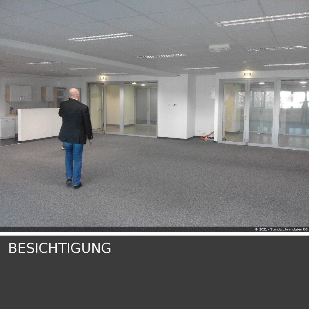 Geräumiger offener Bürobereich mit Teppichboden, integrierter Küchenzeile und großen Fenstern, die viel Tageslicht hereinlassen.