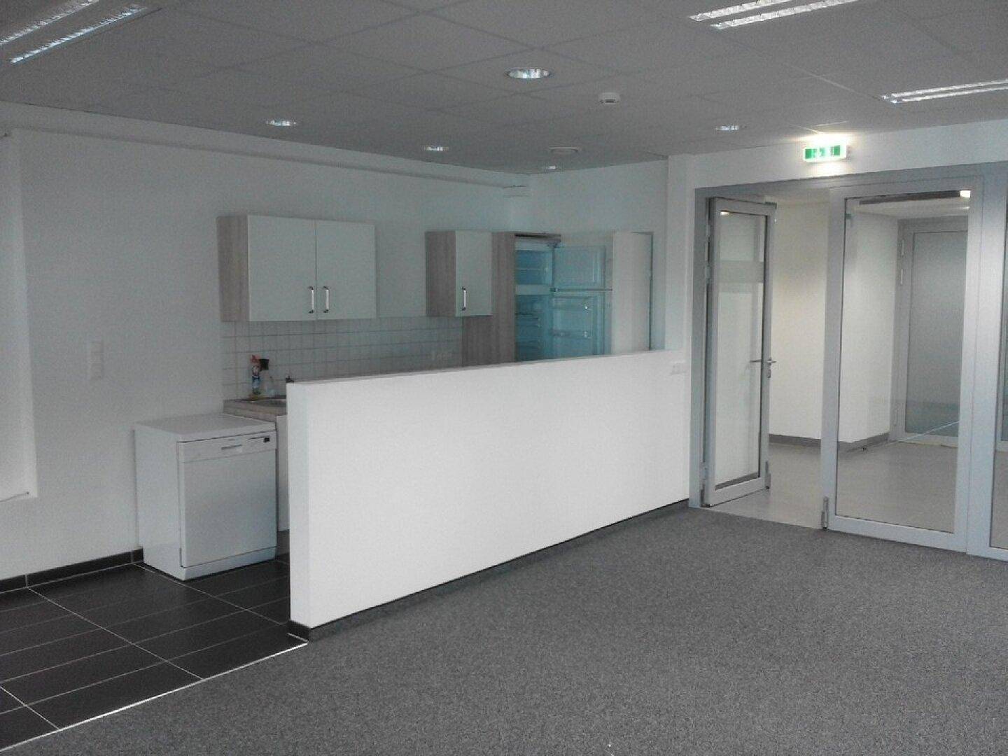 Funktionale Küchenzeile in einem offenen Bürobereich mit weißen Schränken, Spüle und integriertem Kühlschrank.