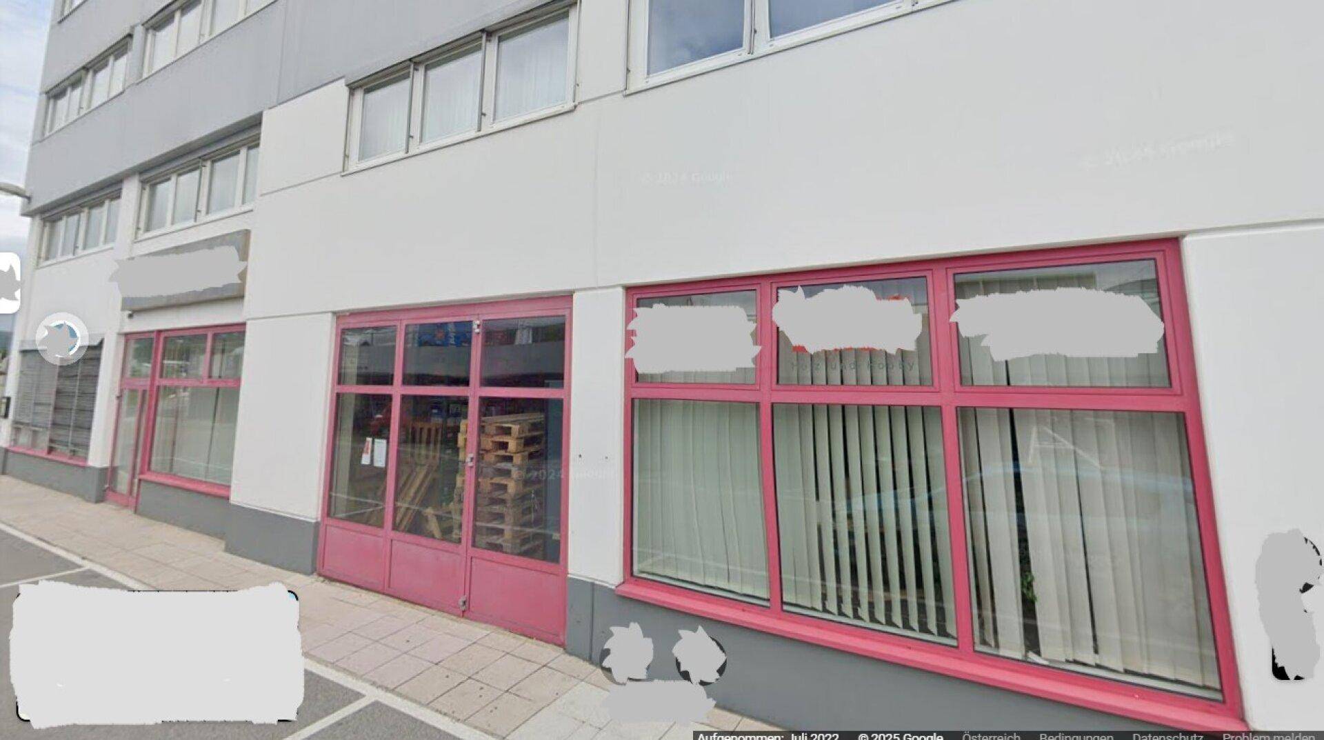 Gebäudeansicht mit weißer Fassade und markanten pinken Fensterrahmen im Erdgeschoss, aufgenommen von der Straße.