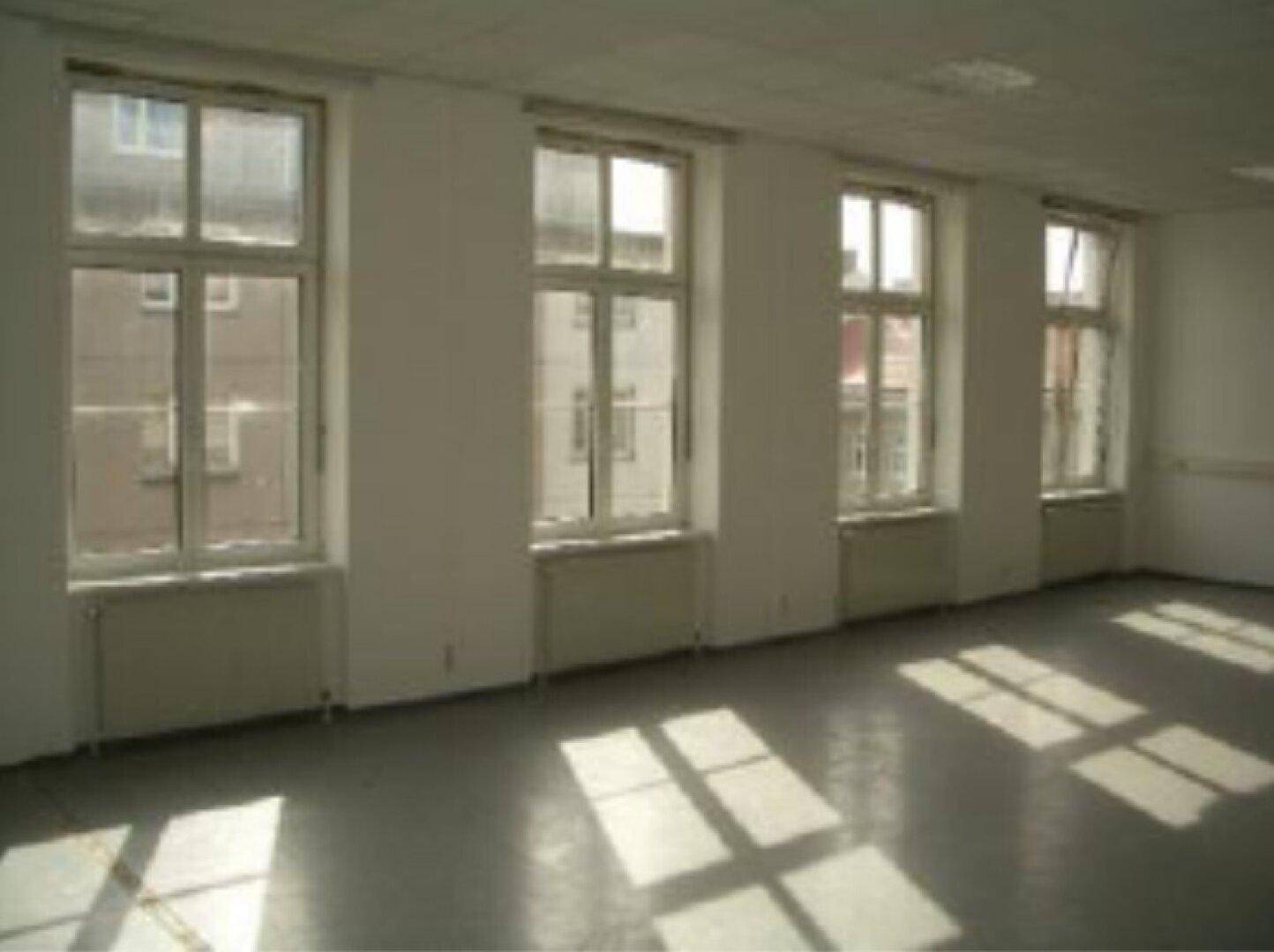 Geräumiger Büroraum mit mehreren Fenstern, die viel Tageslicht hereinlassen und Schatten werfen.