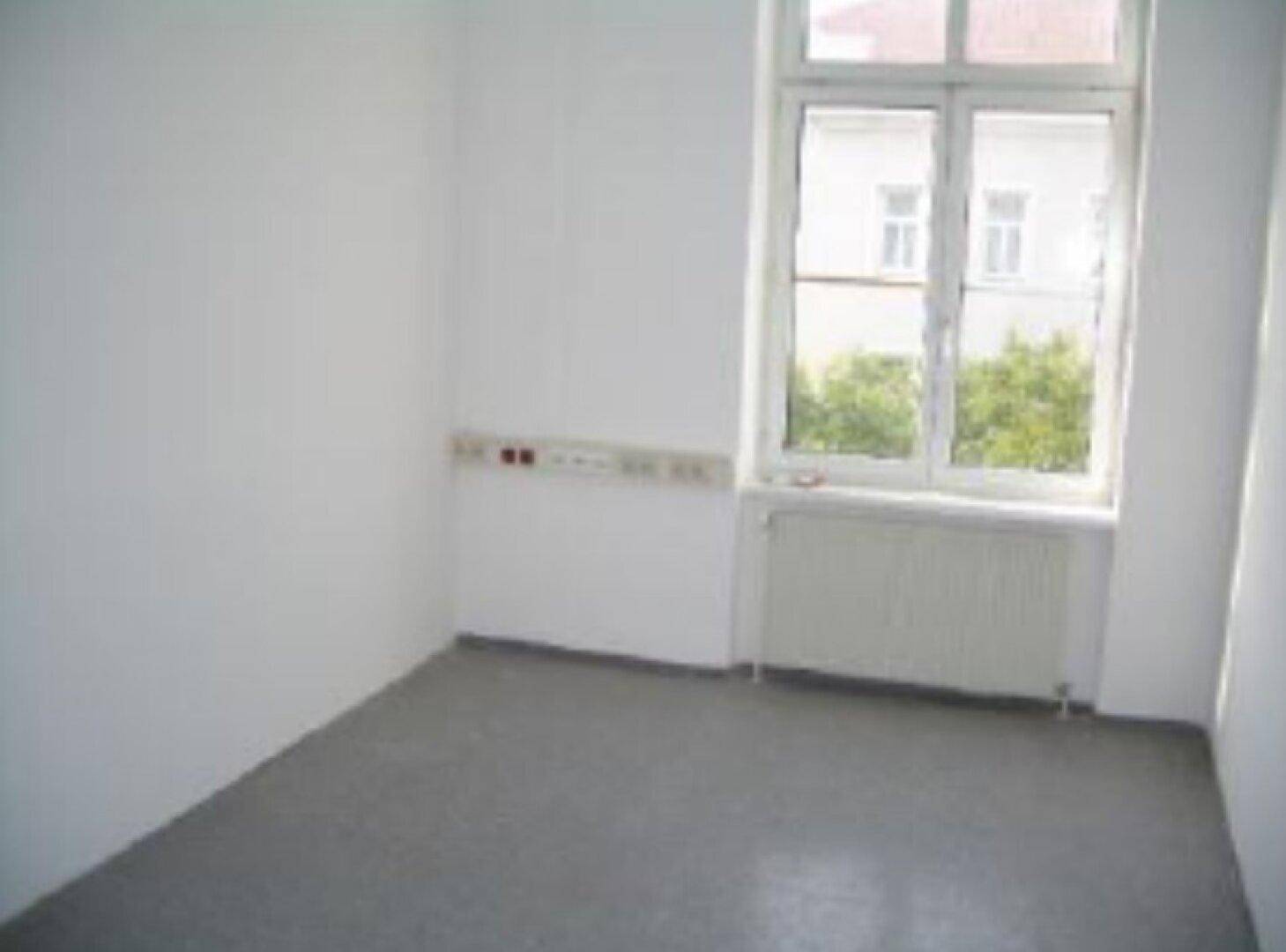 Kleiner, heller Büroraum mit einem großen Fenster, Heizkörper und grauem Bodenbelag.