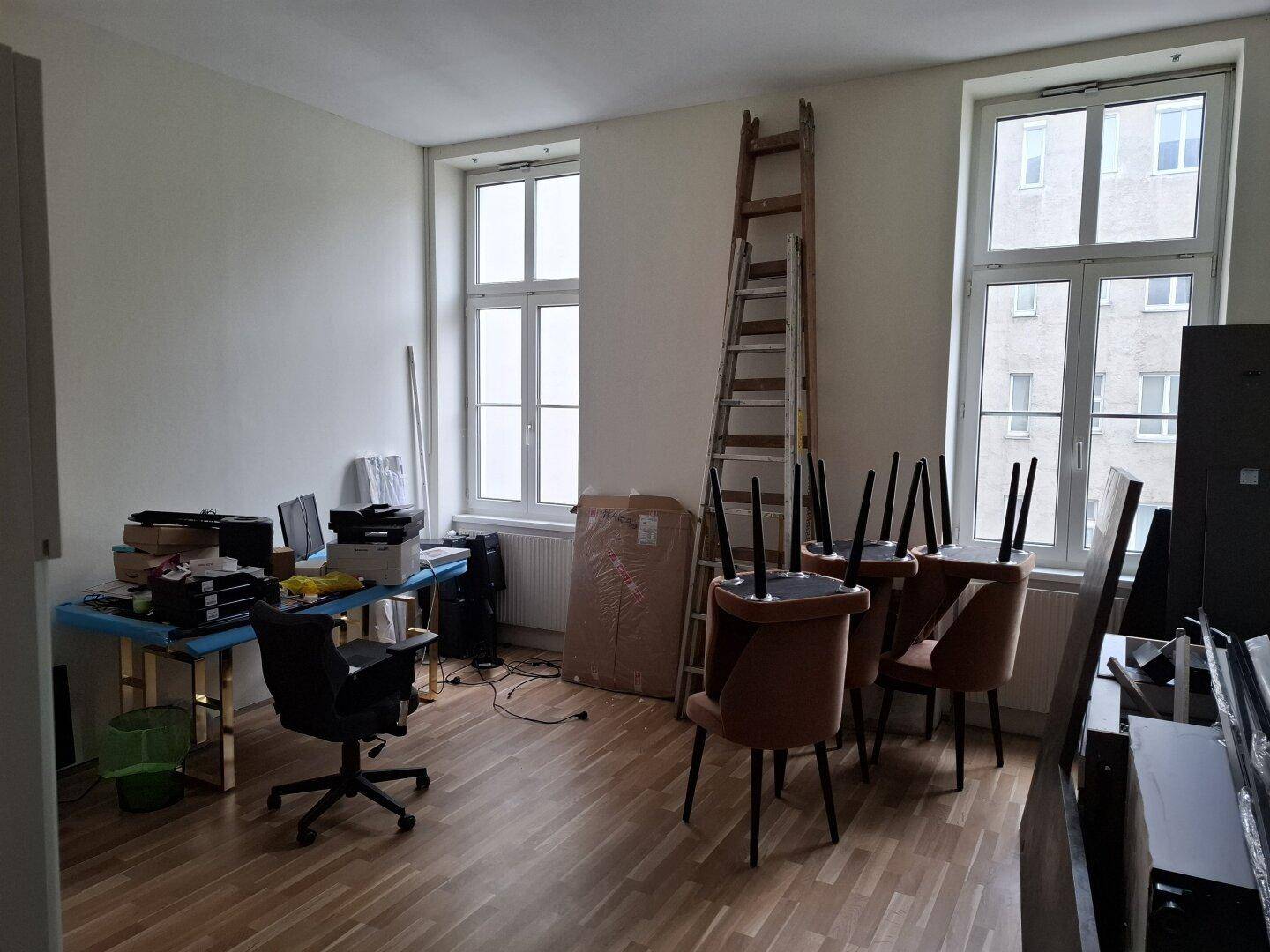 Büroraum mit Holzfußboden, zwei großen Fenstern und verschiedenen Möbeln und Gegenständen.
