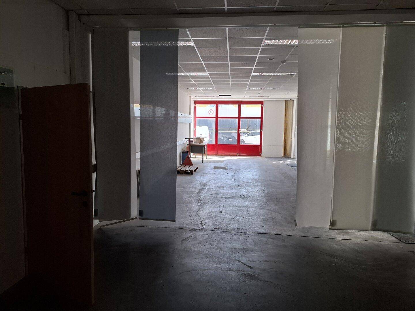 Blick durch eine Öffnung in einen weiteren Gewerbebereich mit Betonboden und markanten roten Türen im Hintergrund.