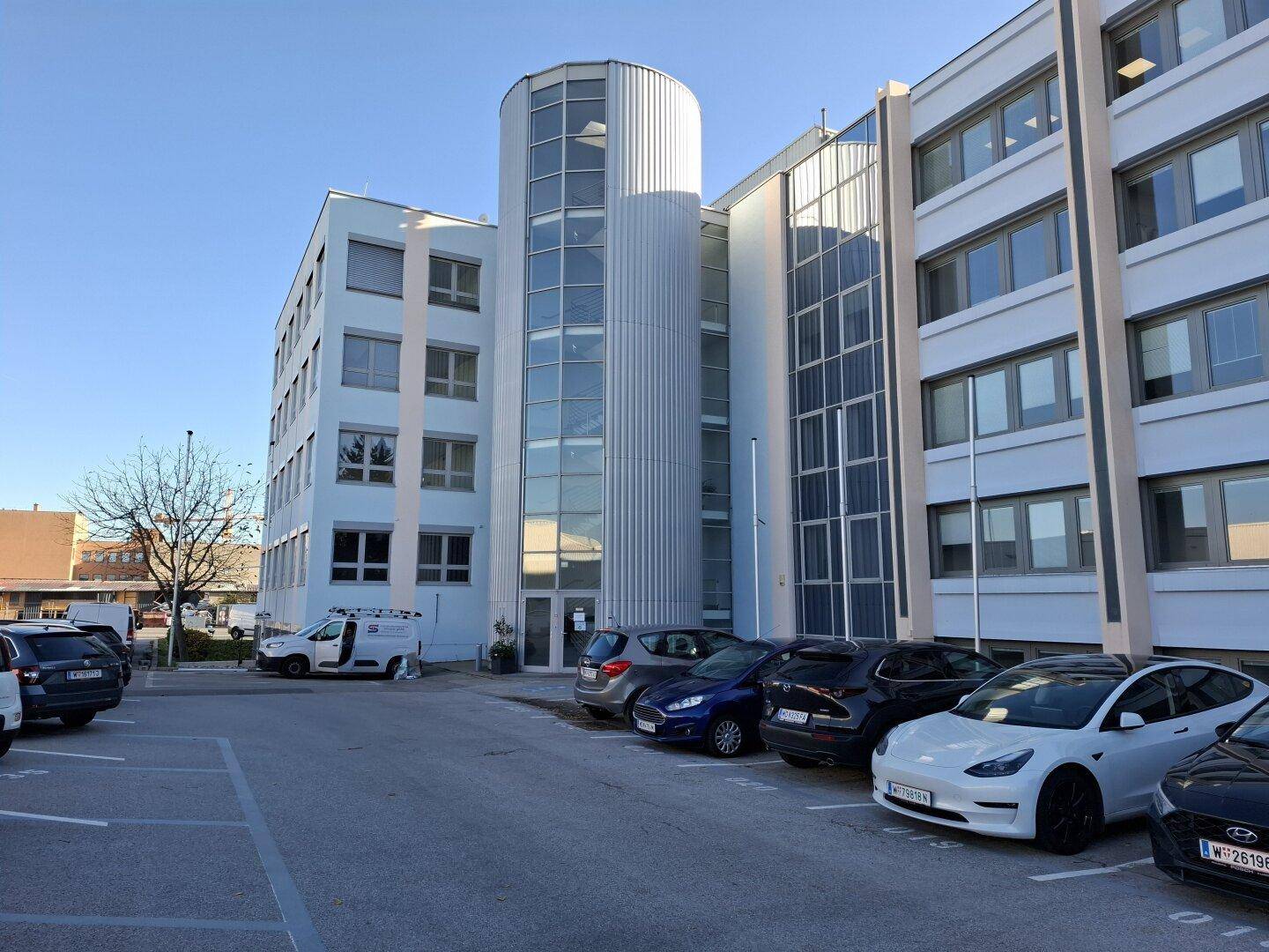 Modernes Bürogebäude mit markanter Glasfassade und zylindrischem Treppenhaus, umgeben von einem Parkplatz.