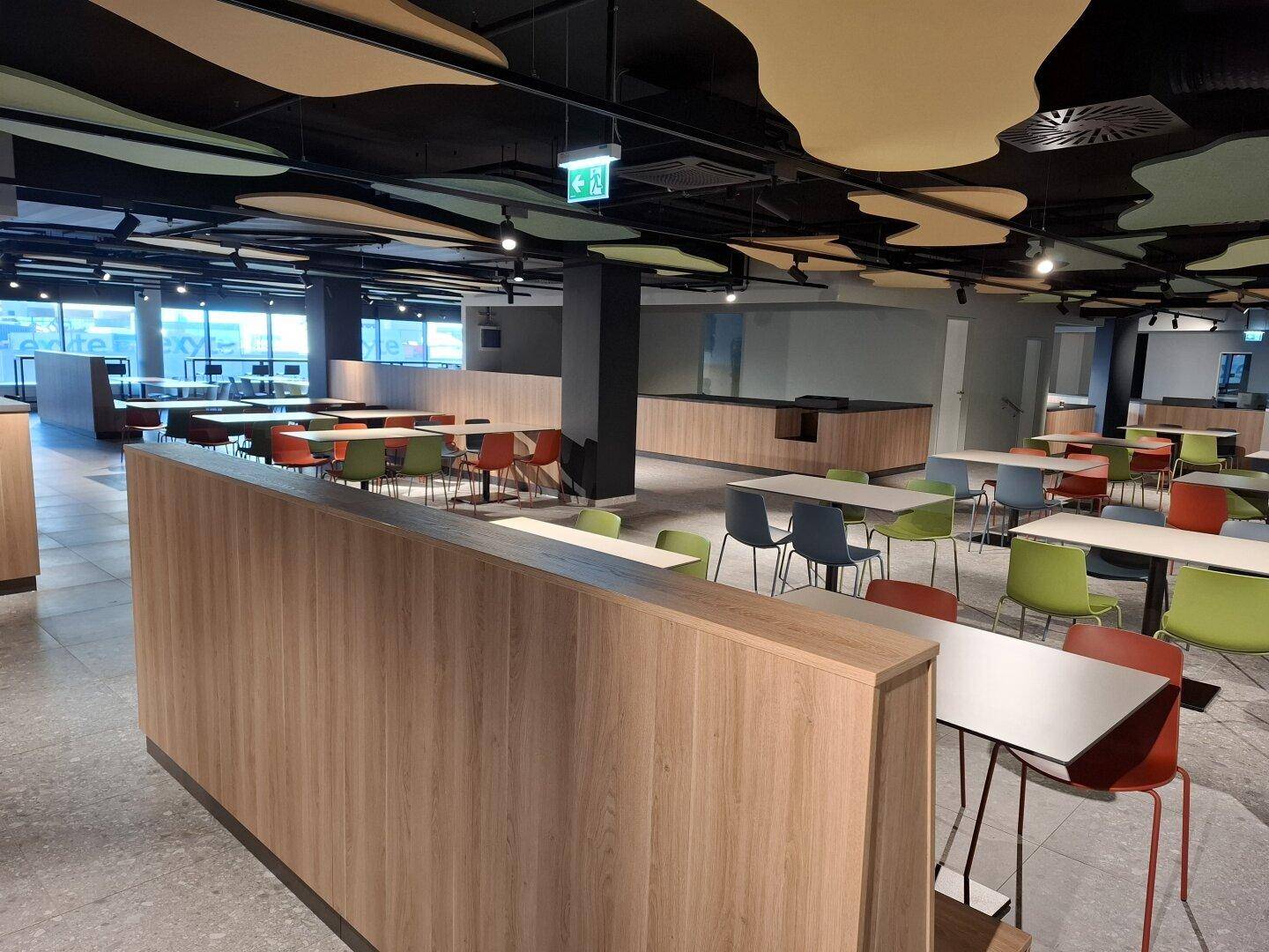 Moderne Cafeteria mit vielen Tischen, bunten Stühlen und einer einzigartigen Deckenverkleidung.