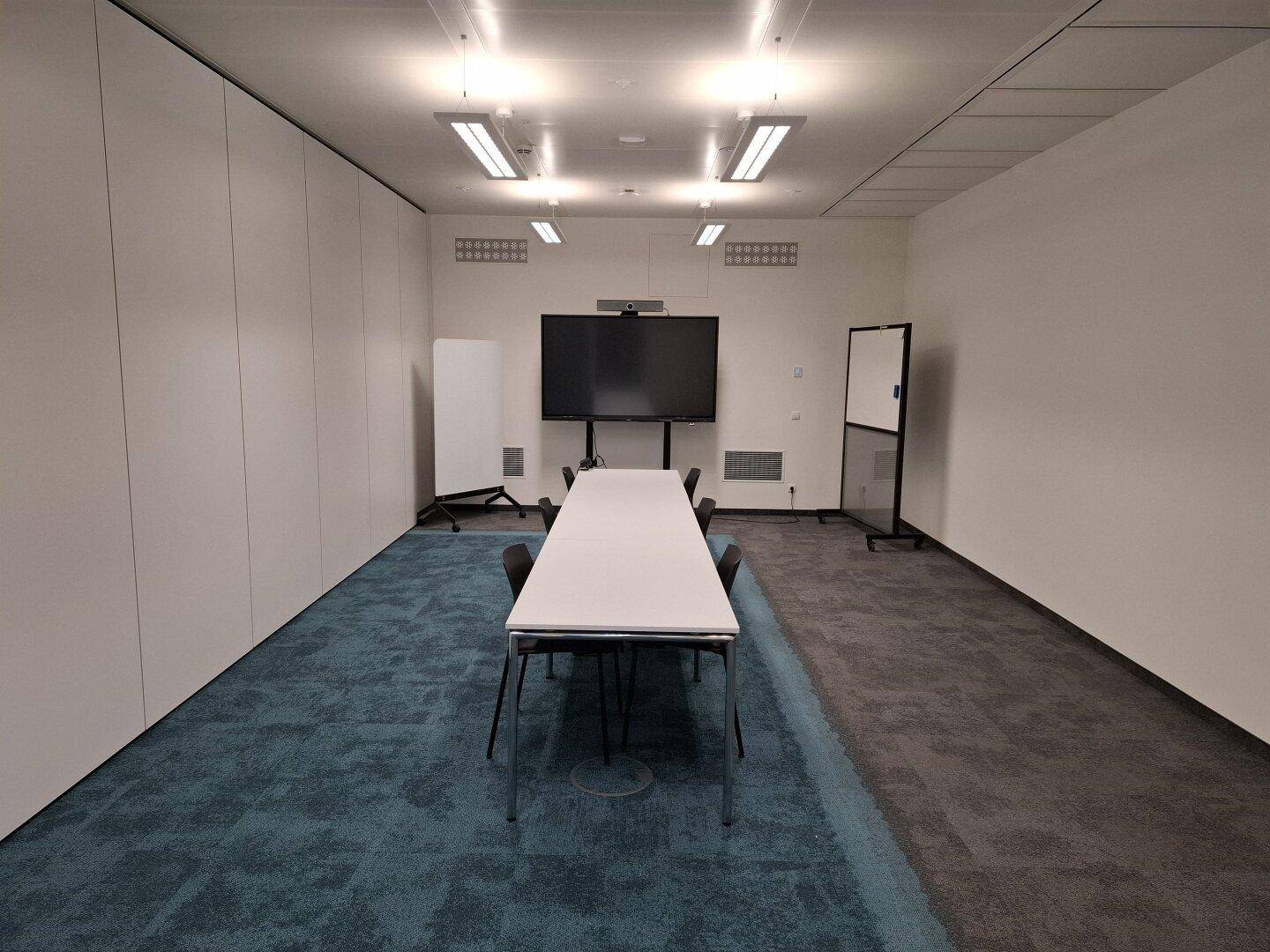 Kleiner Konferenzraum mit langem Tisch, Stühlen, großem Bildschirm und zweifarbigem Teppichboden.