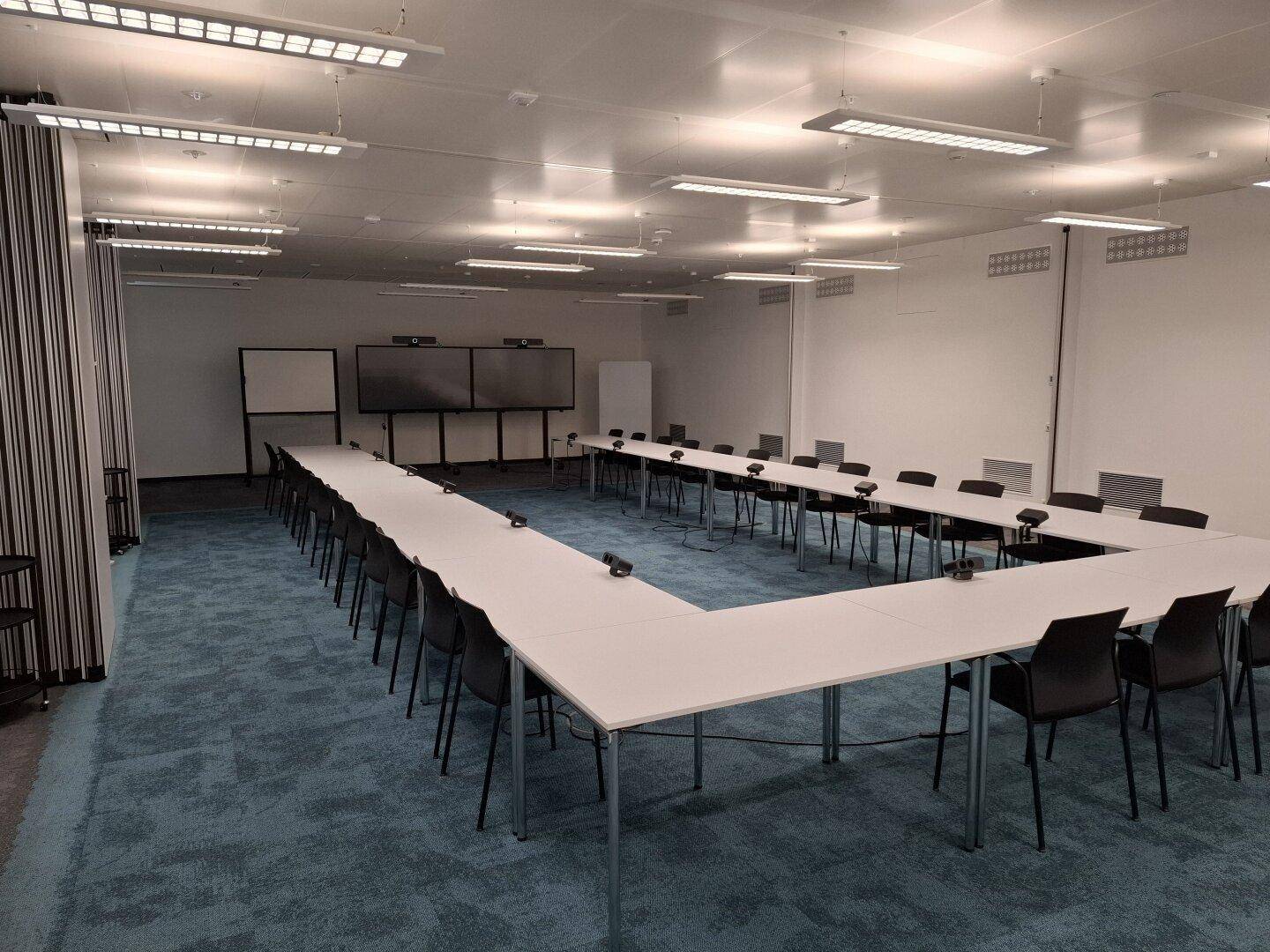 Großer Konferenzraum mit U-förmiger Tischordnung, vielen Stühlen und modernem Teppichboden.