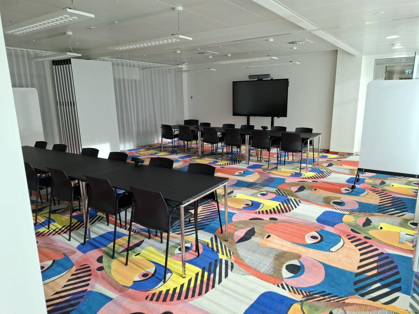 Großer Konferenzraum mit auffälligem, farbenfrohem Teppich, mehreren Tischen und einem großen Bildschirm.