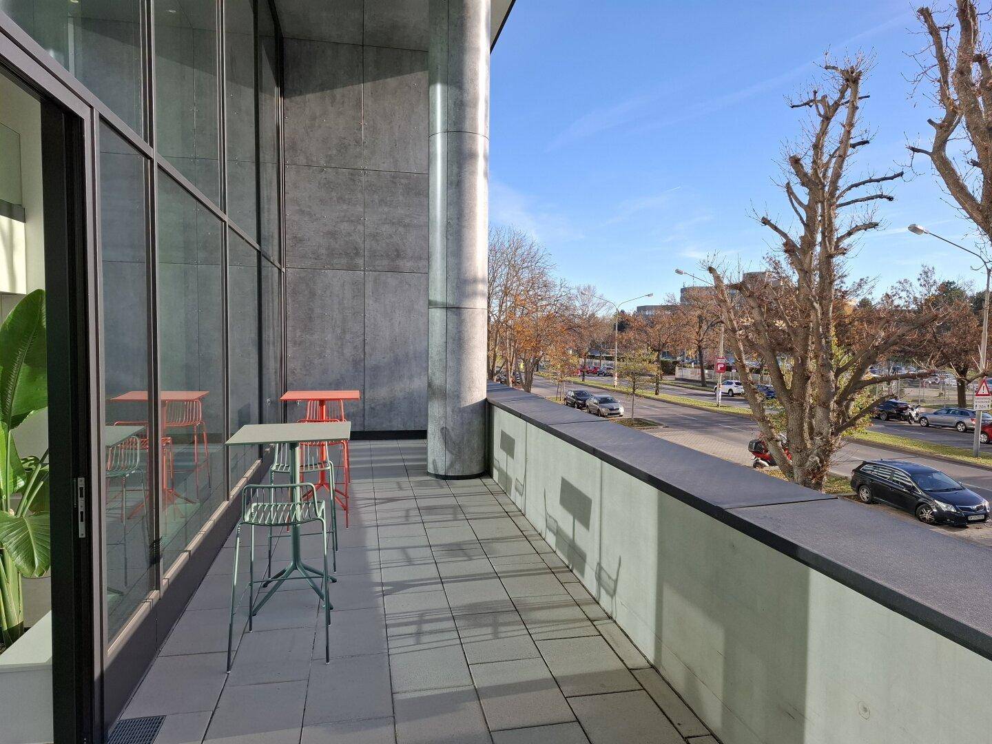 Moderner Balkon mit Fliesenboden, kleinen Tischen und Stühlen, mit Blick auf die Straße.